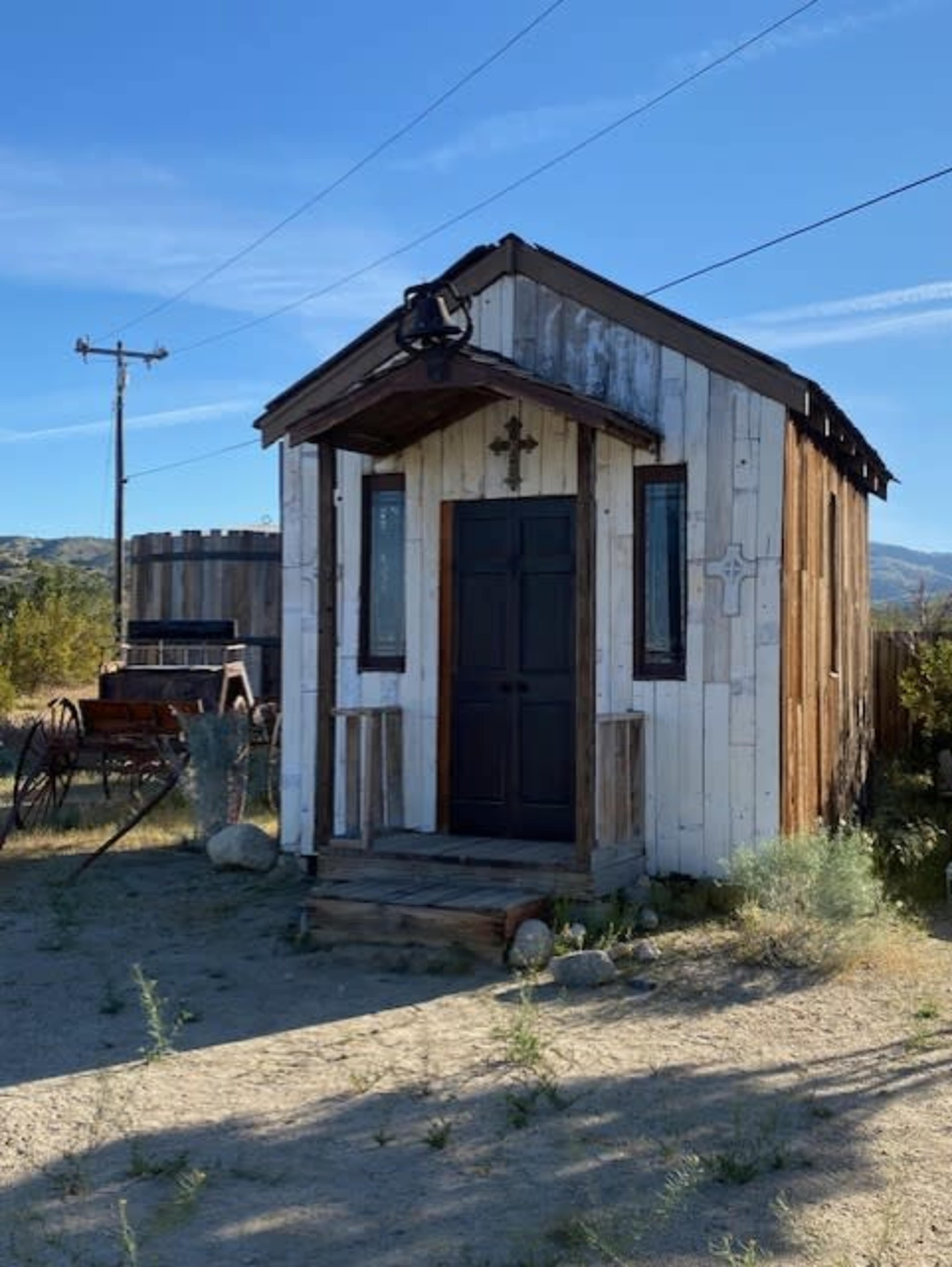 The image shows a weathered wooden shed with a dark door, located in a dust-covered area with sparse vegetation and a distant wagon.
