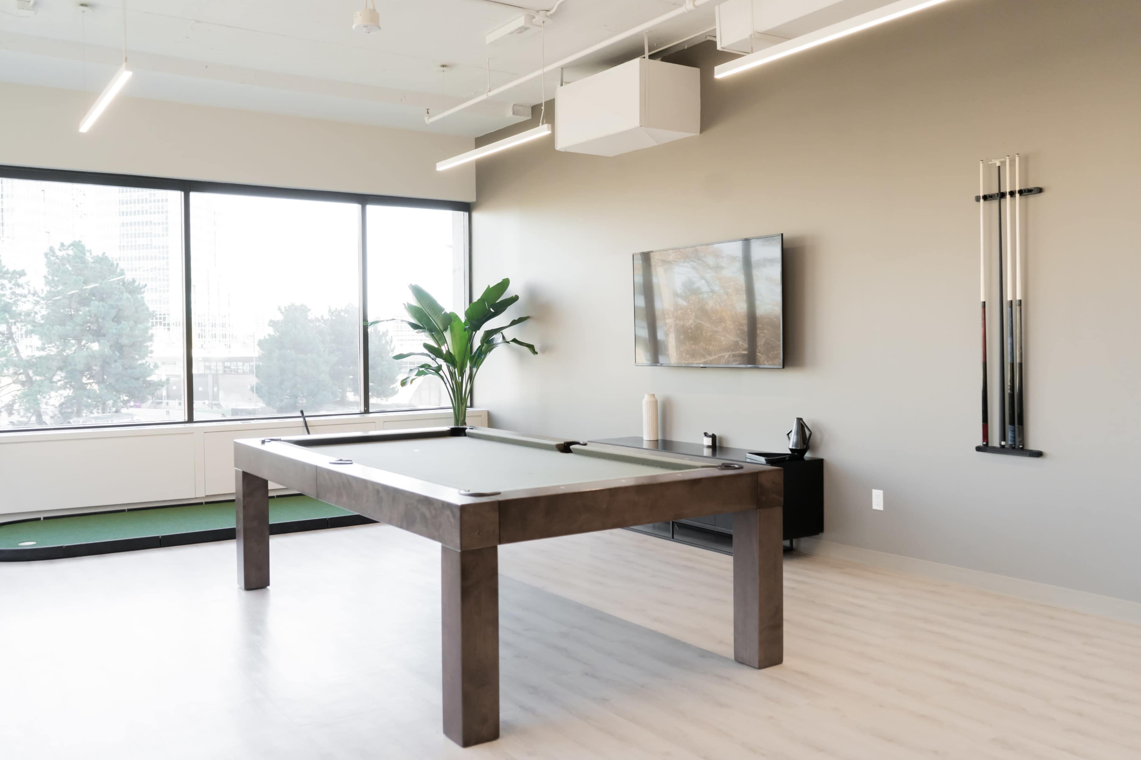 A modern game room features a pool table, a flat-screen TV mounted on the wall, and large windows overlooking a cityscape.