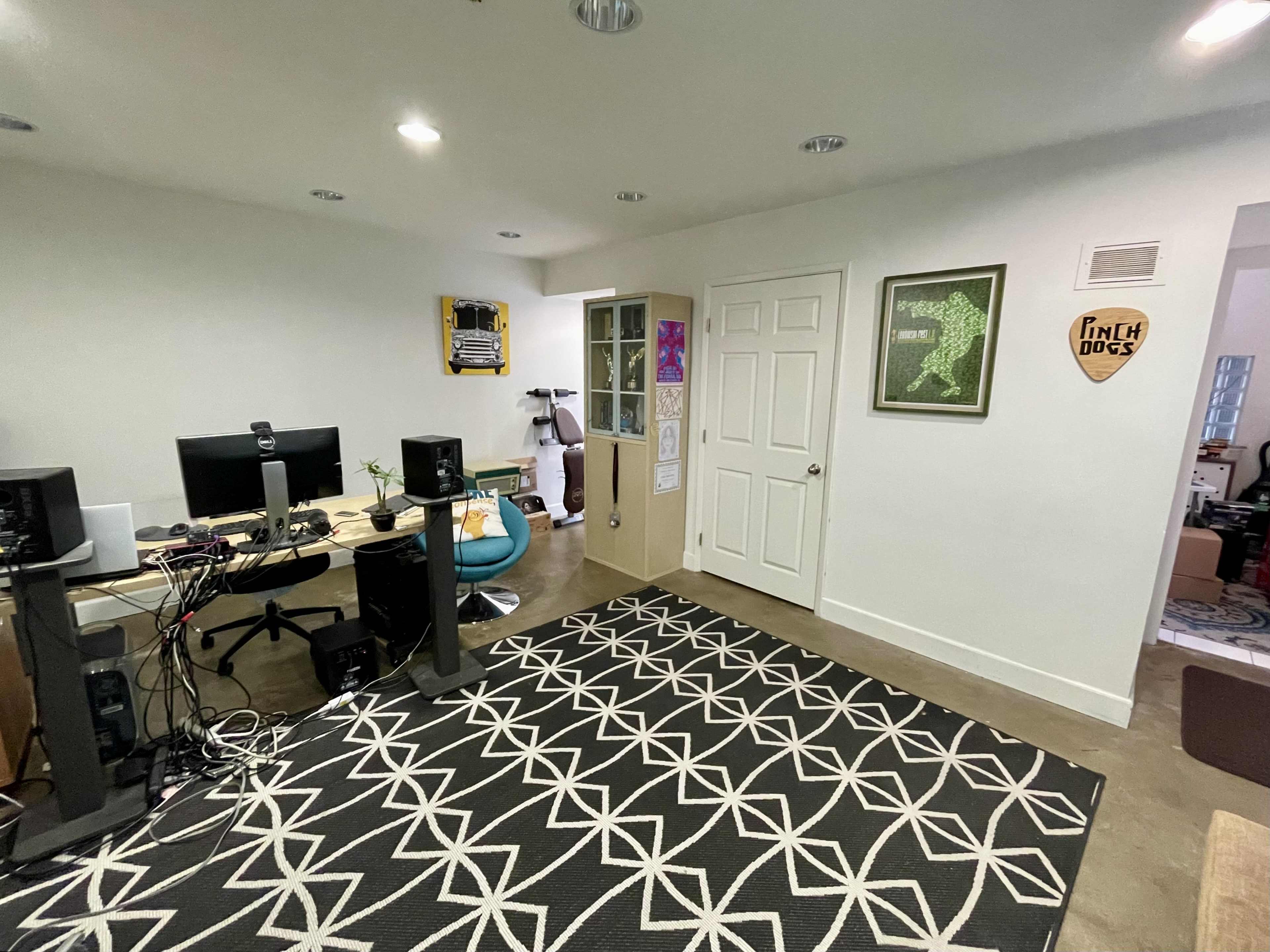 The image shows a spacious room with two computer desks, a large area rug, and a door leading to another space, featuring decorative wall art.