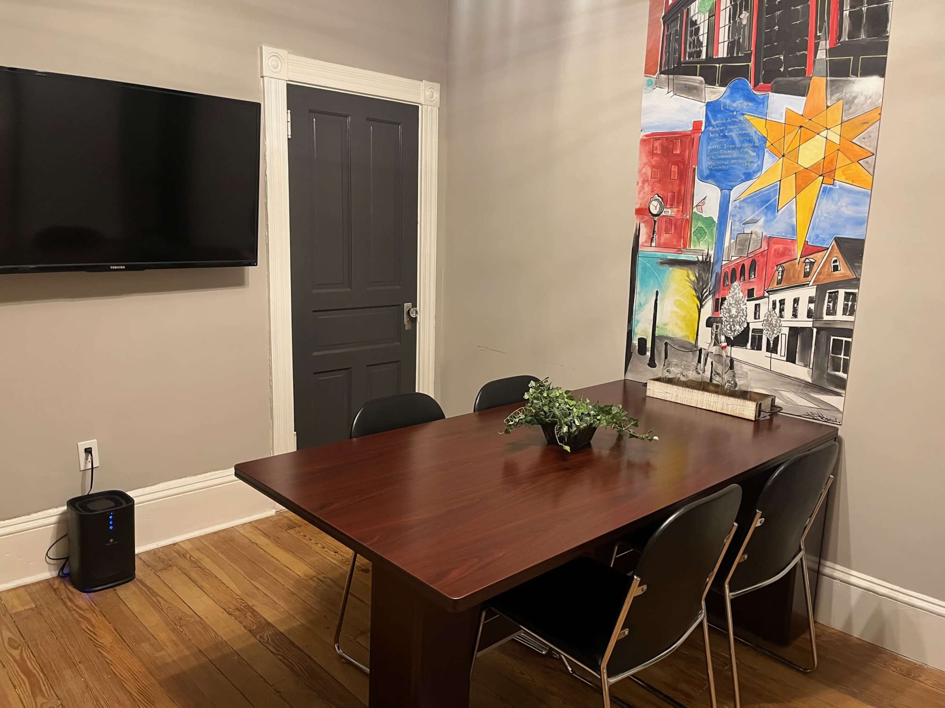 A meeting room features a wooden table with four black chairs, a television mounted on the wall, and a colorful mural in the background.