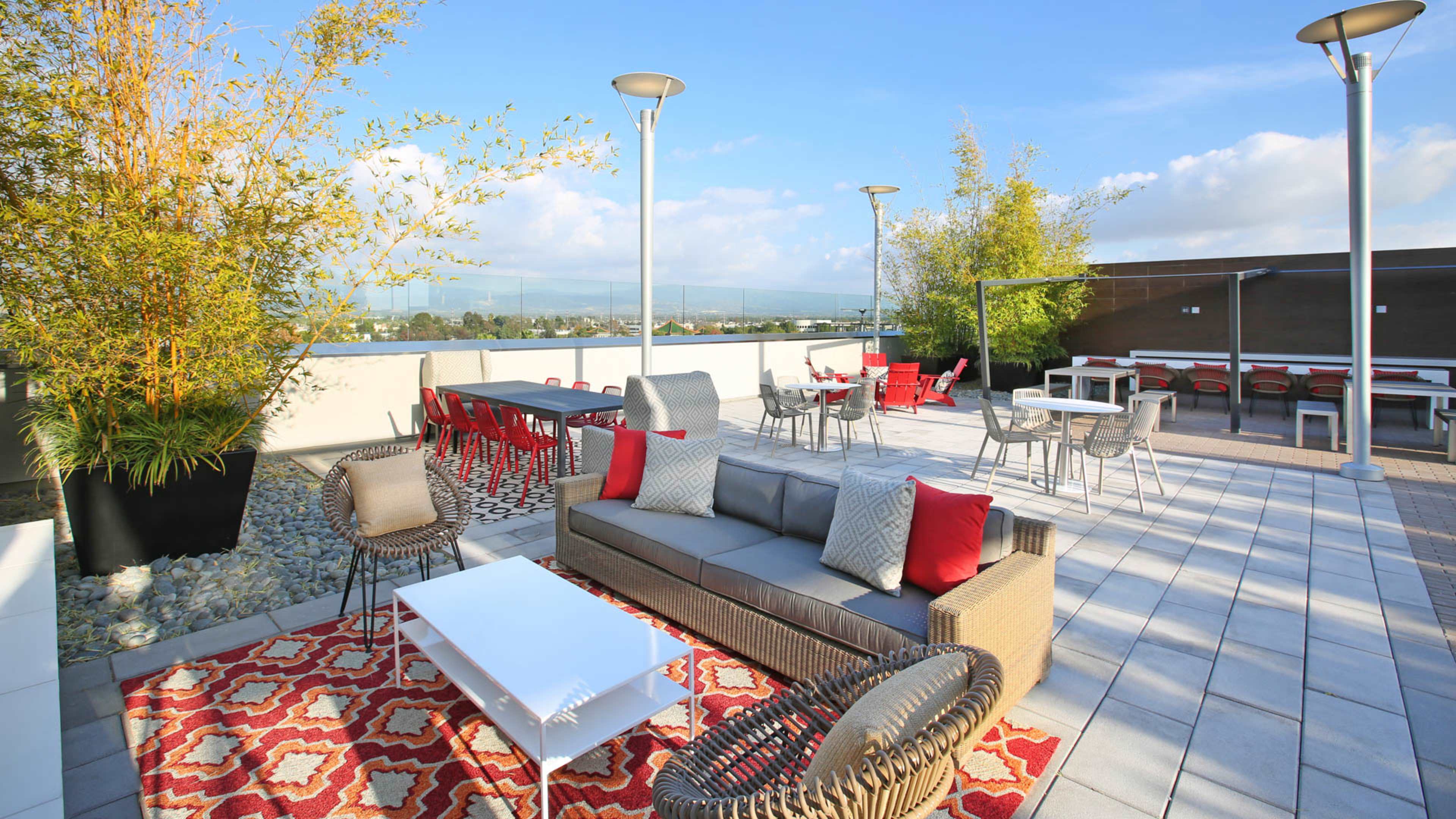 A rooftop terrace features modern seating arrangements, a table, and decorative planters against a clear blue sky.