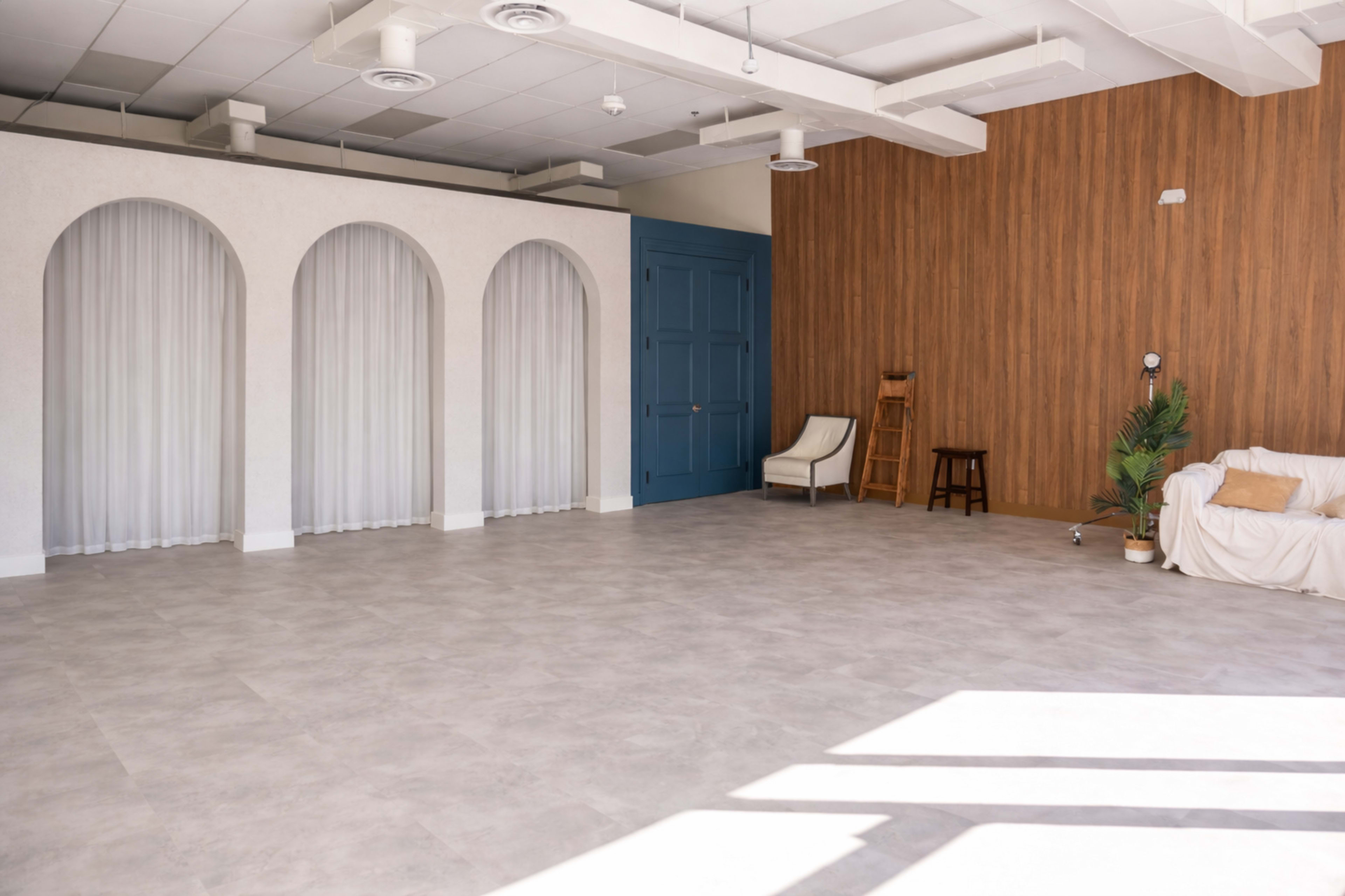 The image shows a spacious, minimalist indoor area featuring arched walls, a blue door, wooden paneling, and a few pieces of furniture.