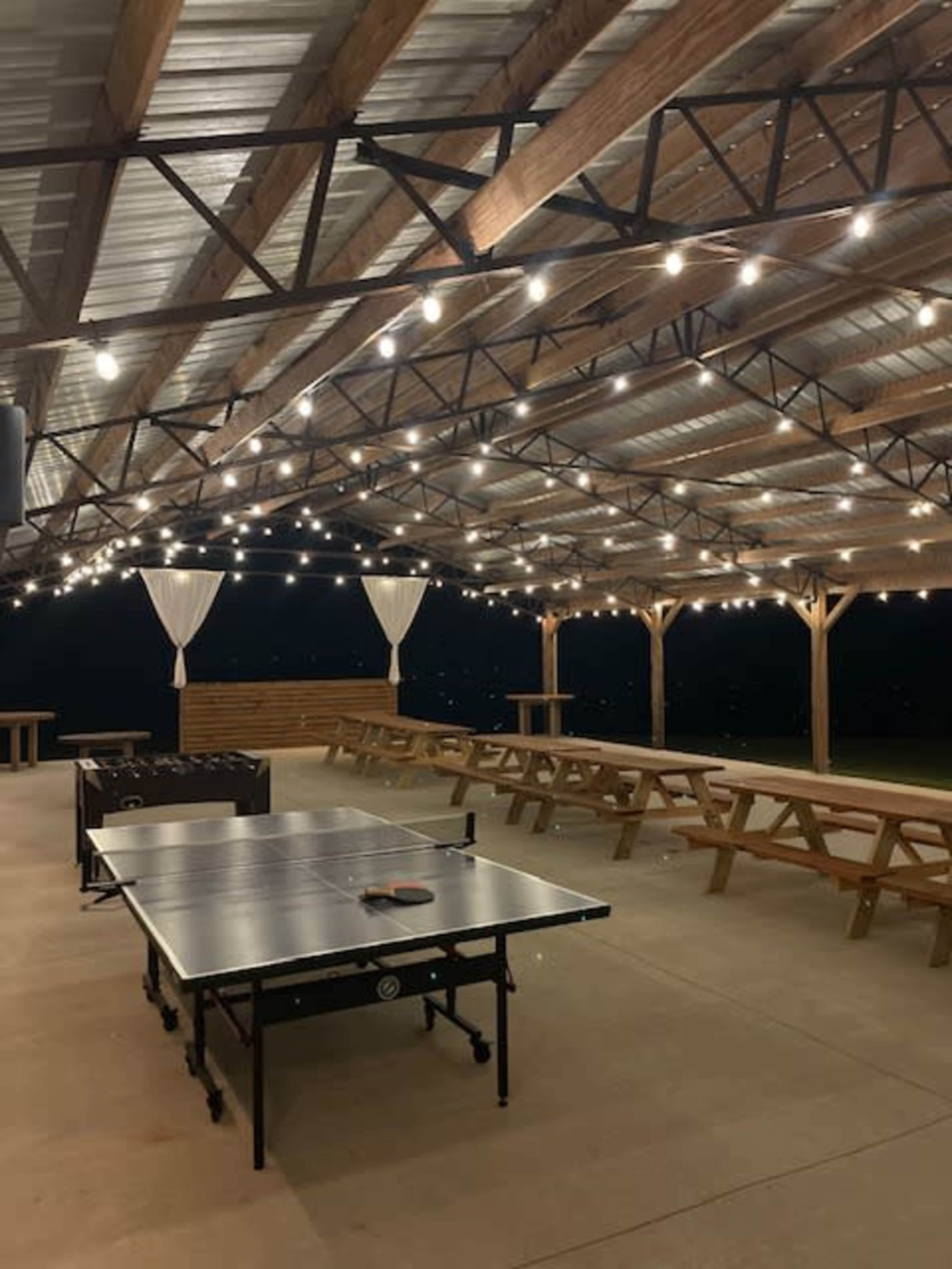 An outdoor pavilion with string lights above and a ping pong table in the foreground, surrounded by wooden picnic tables.