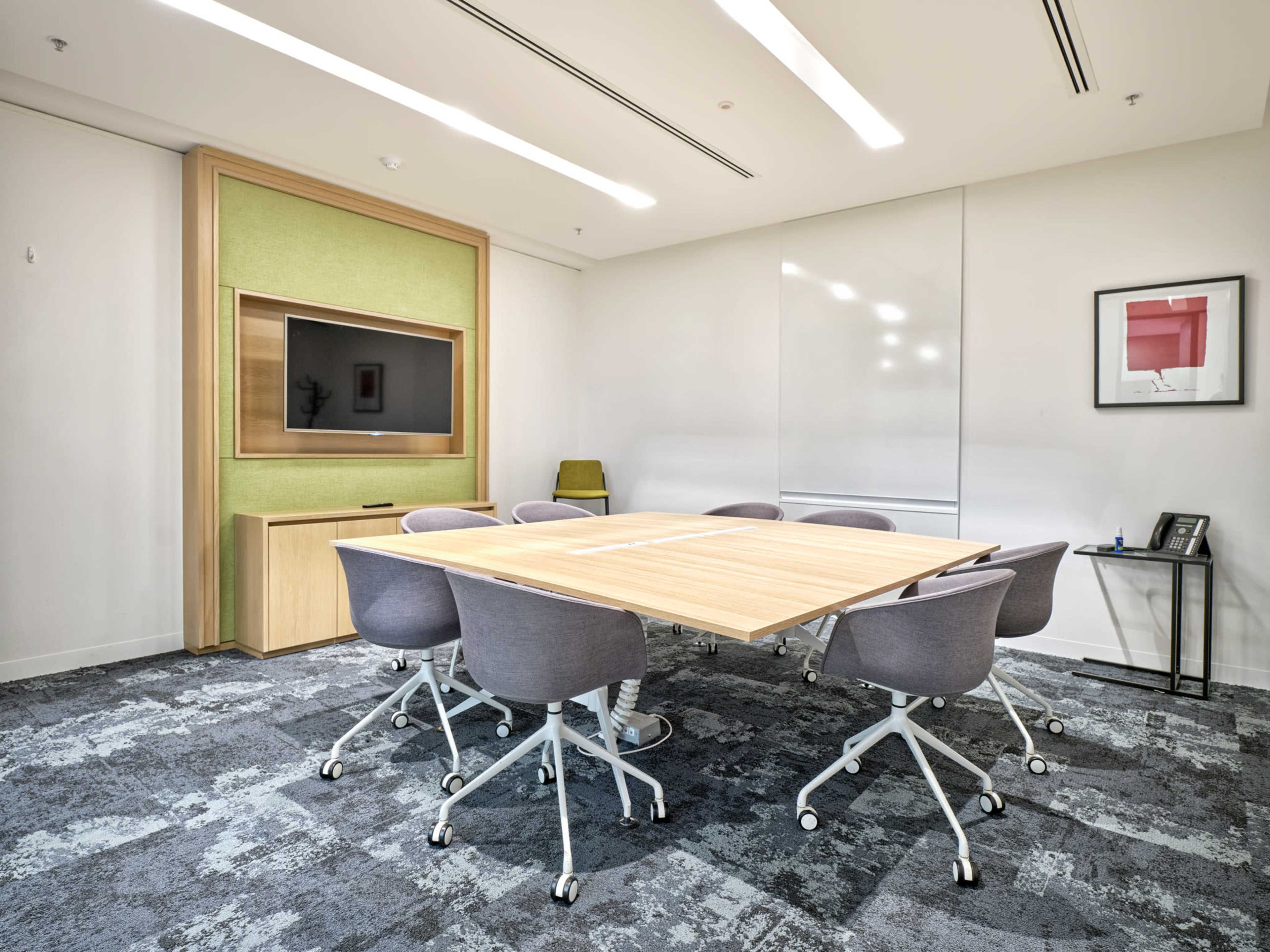 A modern conference room features a large wooden table surrounded by gray chairs and a wall-mounted TV.