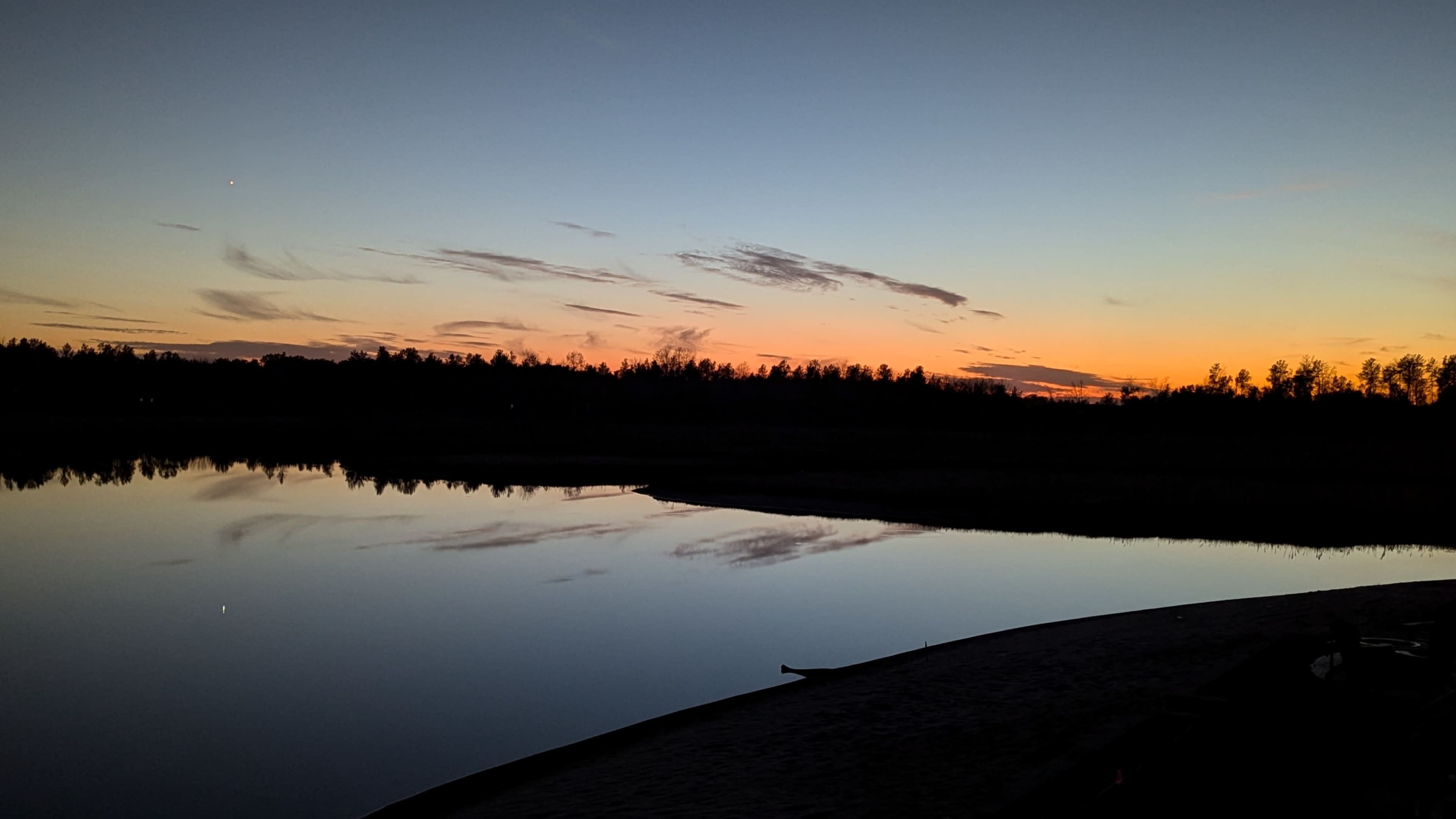 The image shows a tranquil sunset reflecting on a calm body of water, with silhouettes of trees along the horizon.