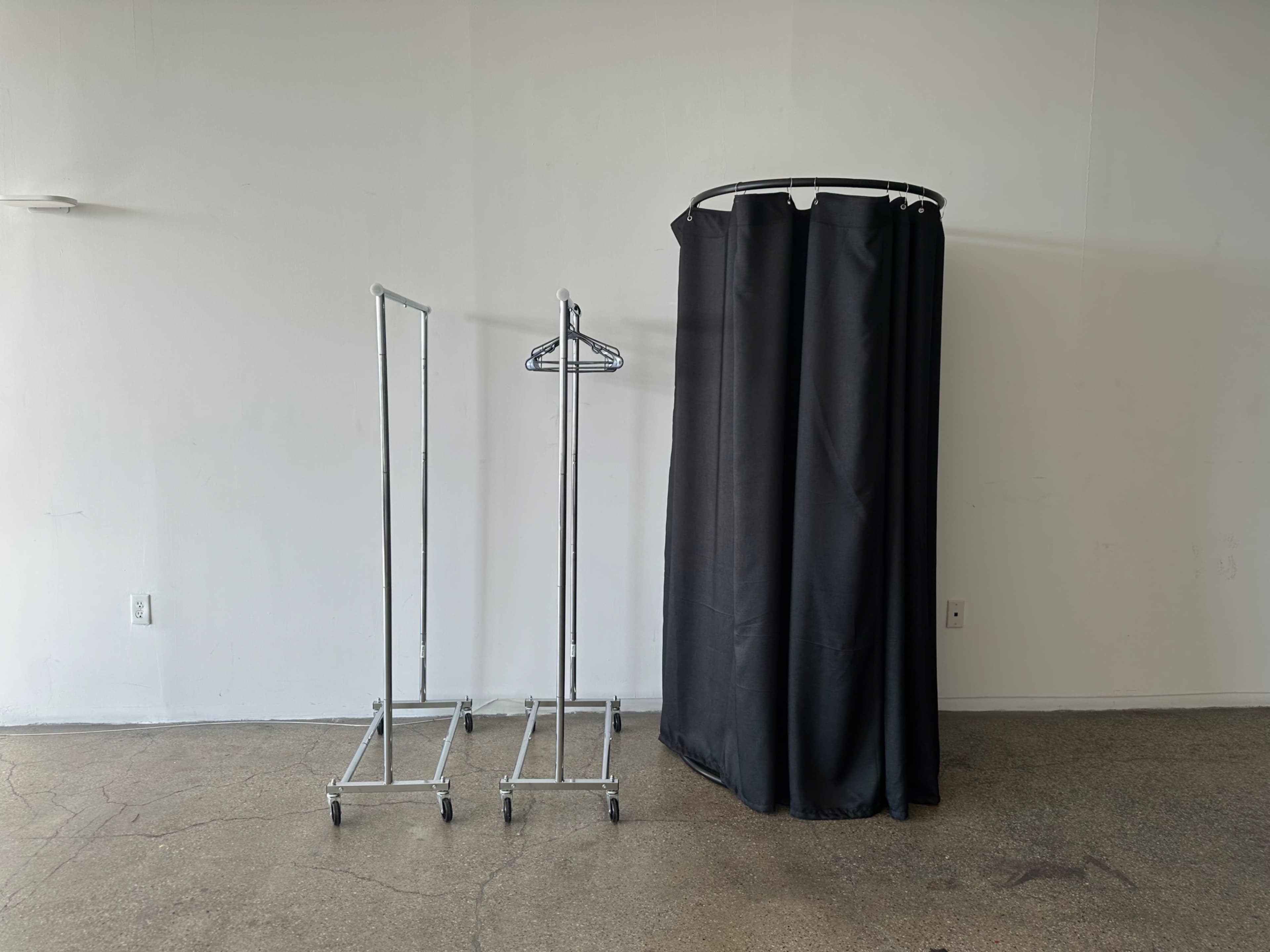 The image shows two clothing racks and a black fabric curtain in an empty room with a concrete floor.