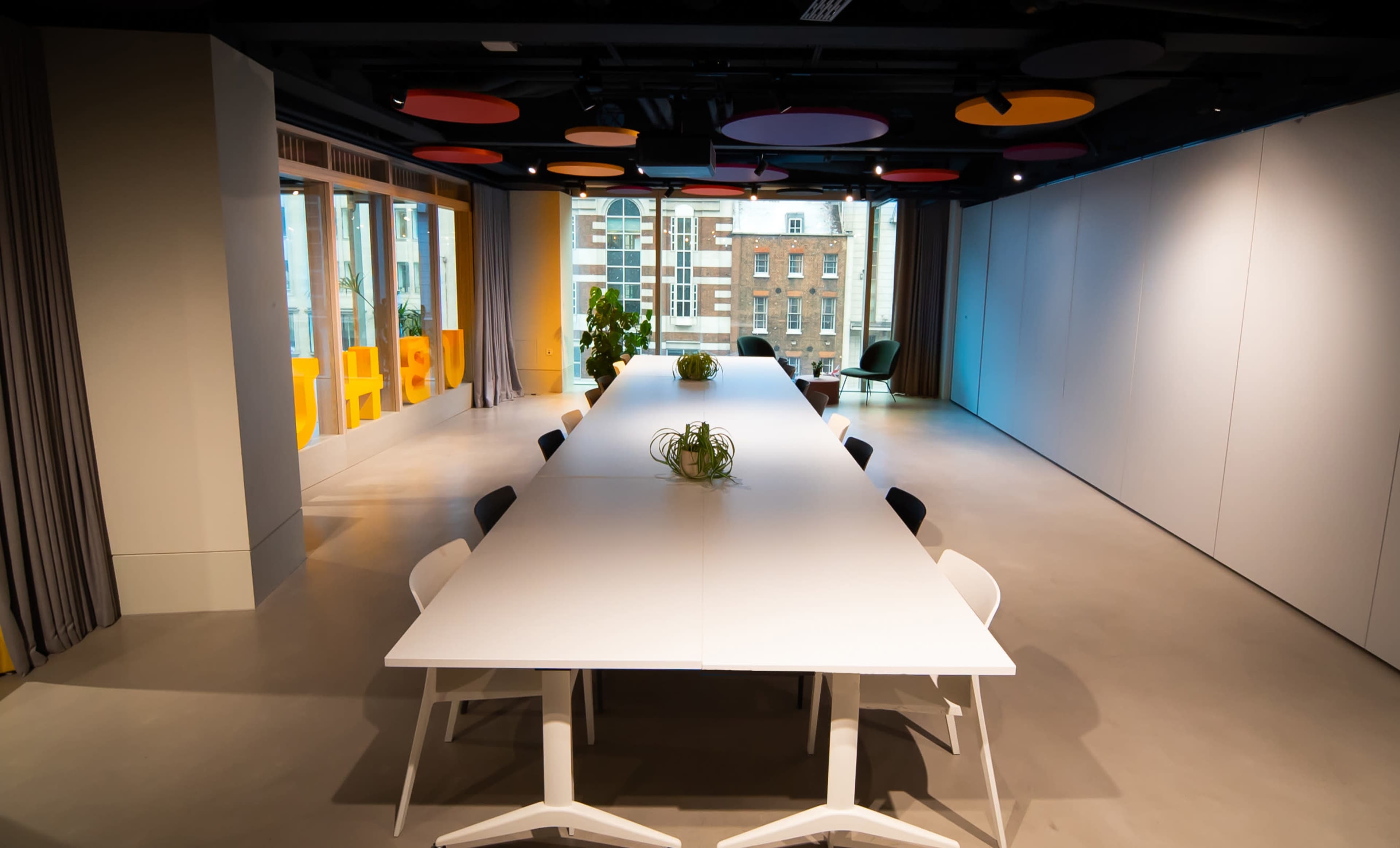 A long, white conference table is centered in a modern meeting room with large windows, colorful ceiling panels, and potted plants.