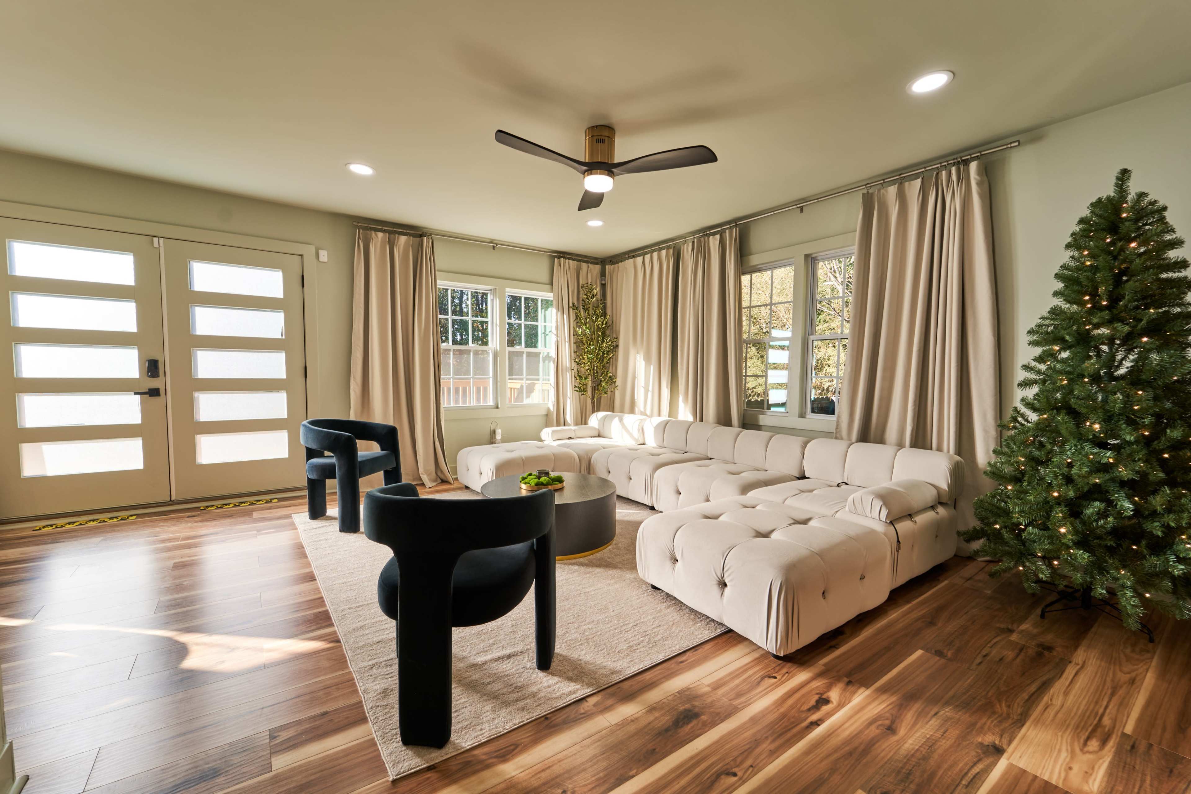 A spacious living room with a light-colored tufted sofa, a round black coffee table, two black chairs, large windows, and a Christmas tree in the corner.