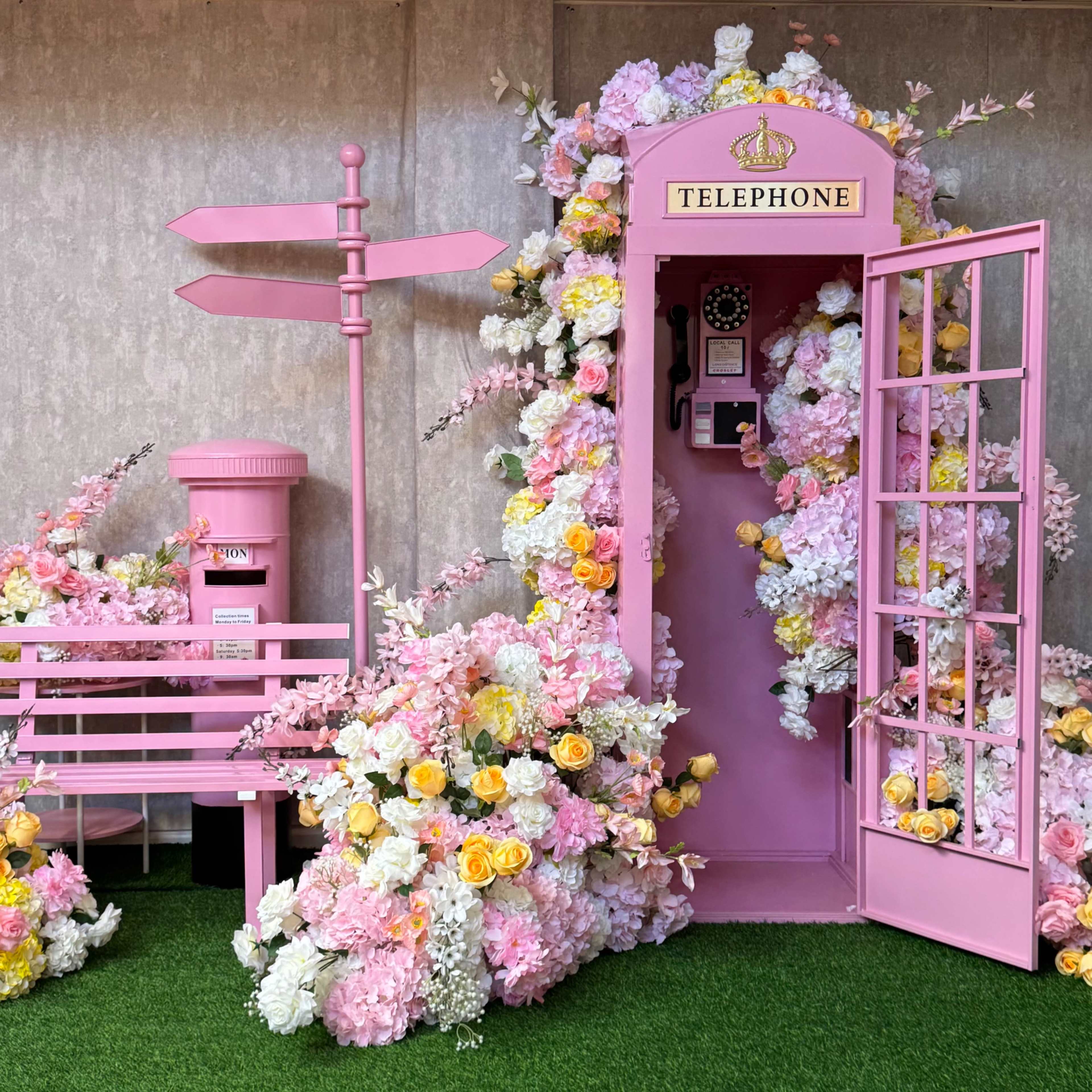The scene features a pink telephone booth surrounded by a vibrant arrangement of flowers, with a matching pink bench and a directional sign nearby.