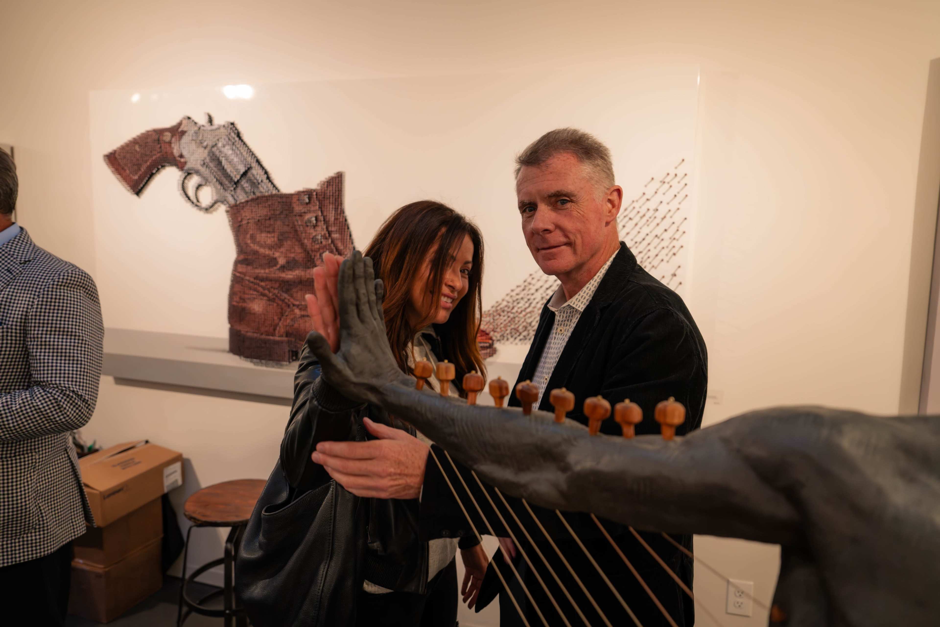 Two people stand in an art gallery, with a sculpture of a hand playing a stringed instrument and artwork featuring a gun and a leather holster in the background.
