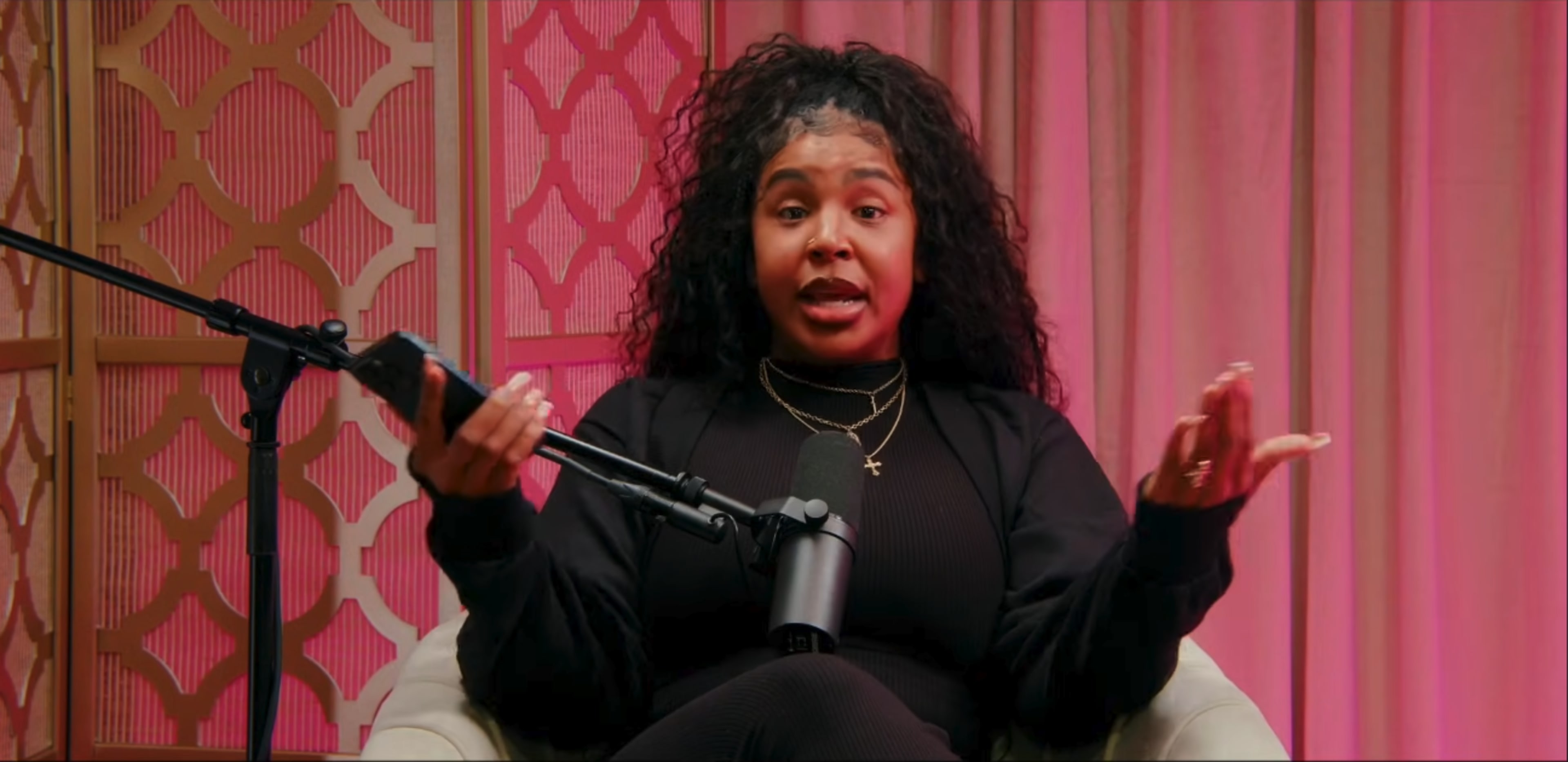 A person with curly hair sits on a sofa, holding a phone and gesturing while speaking, with a microphone in front of them and a decorative backdrop.