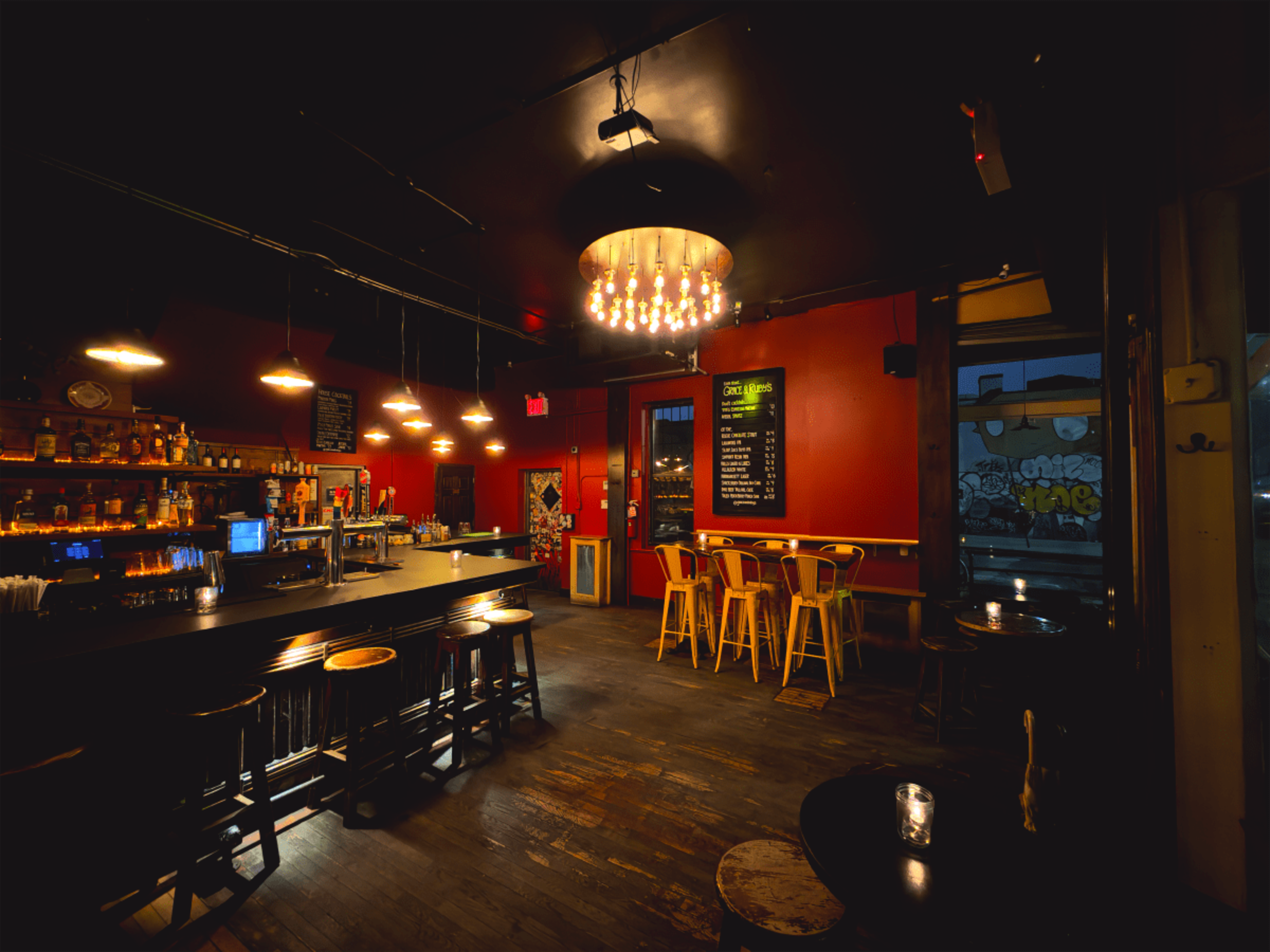 The image shows a dimly lit bar featuring a long counter with stools, a wall-mounted drink menu, and a chandelier, with graffiti visible through a window.