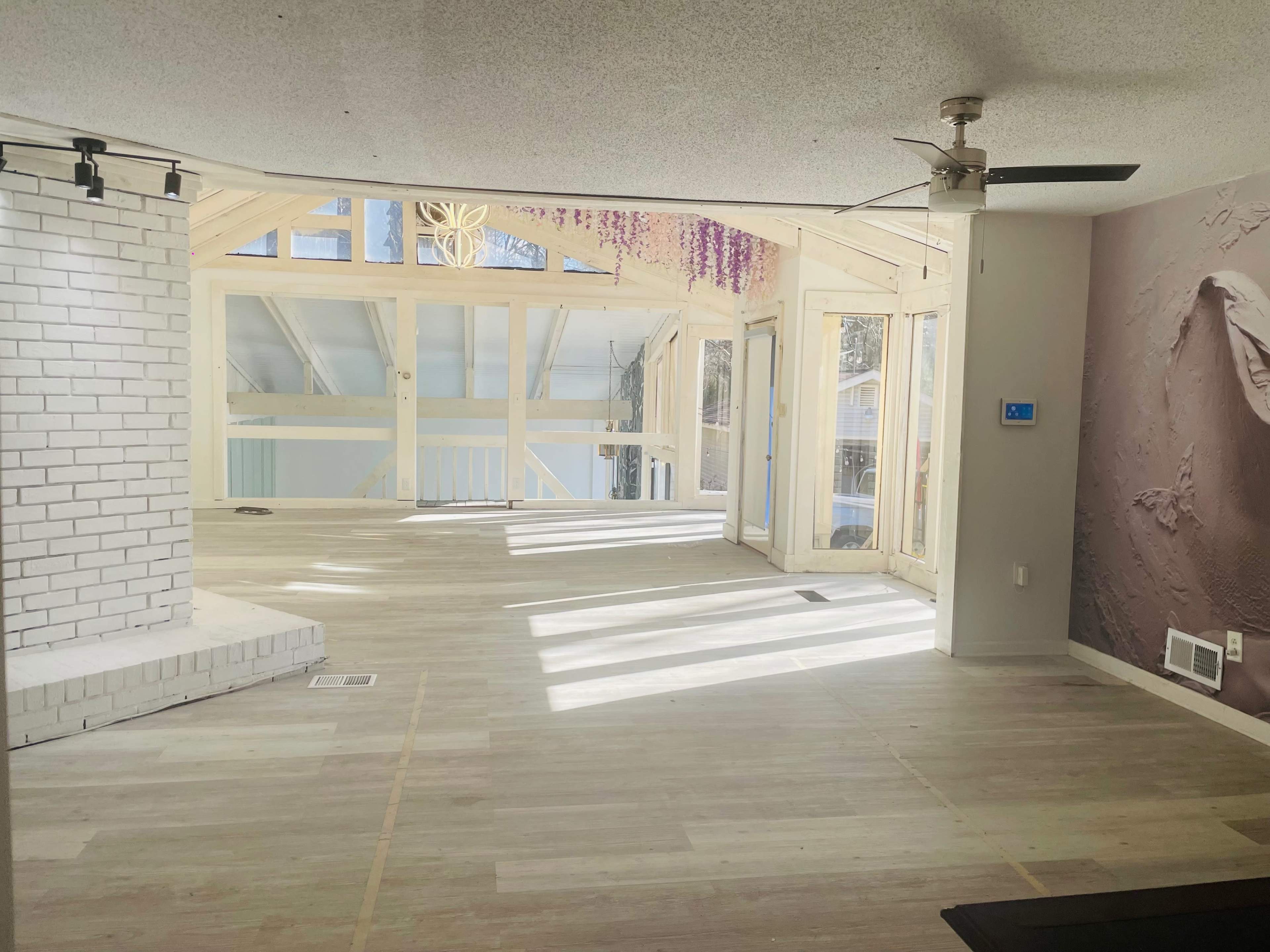 The image shows a spacious room with wood flooring, large windows letting in natural light, and wooden framing around the walls.