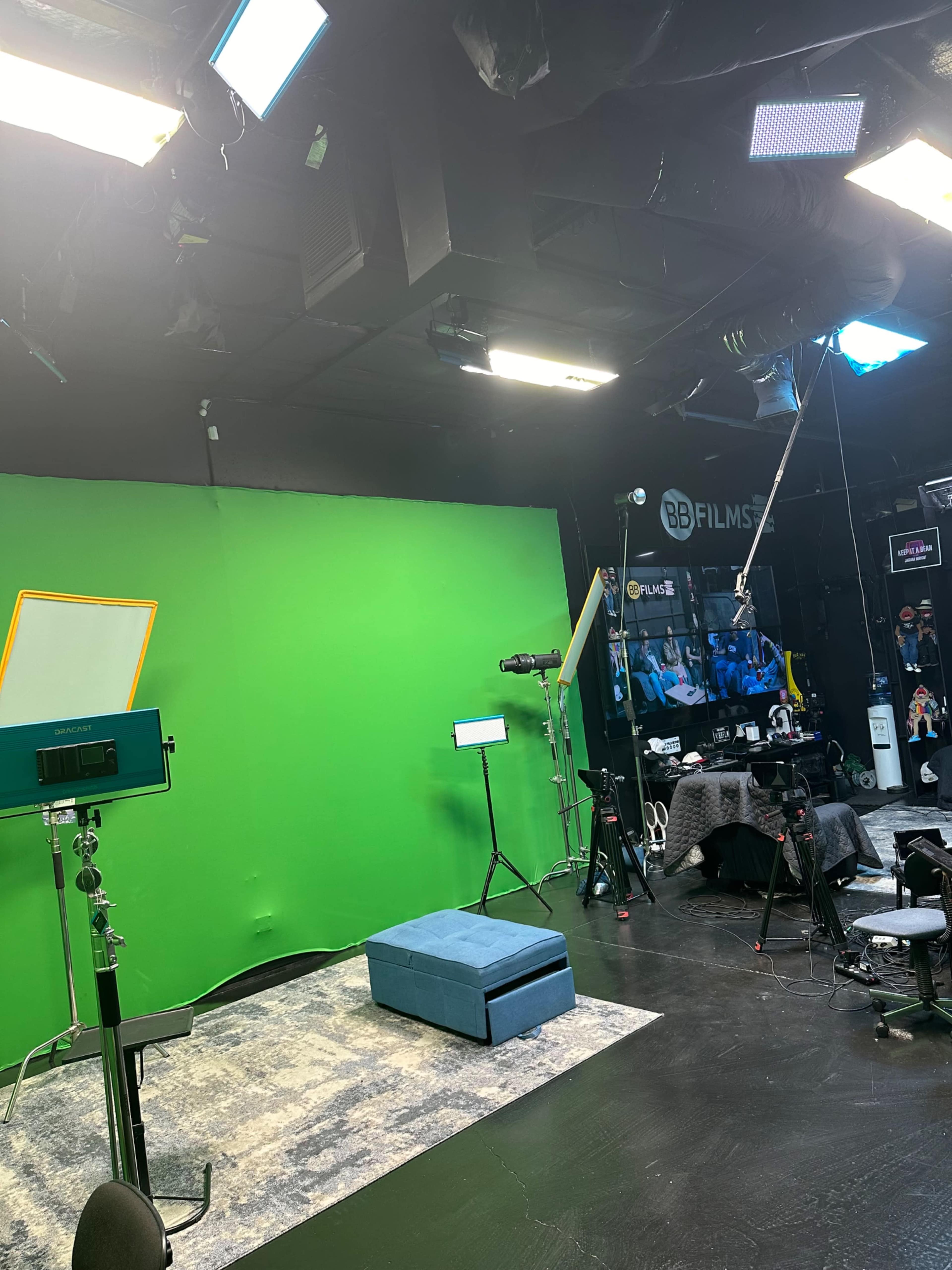 The image shows a studio setup with a green screen, various lighting equipment, cameras, and a blue ottoman on the floor.