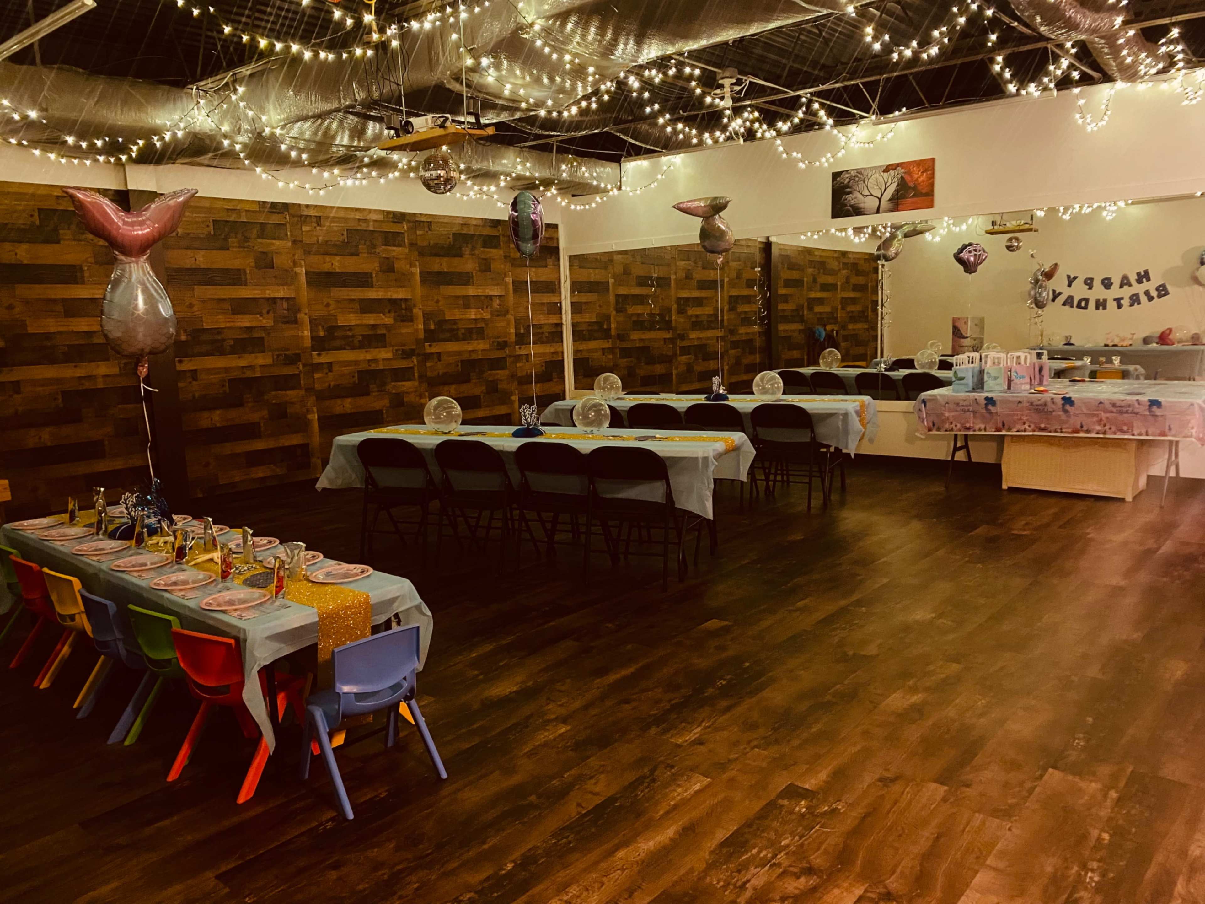A decorated party space with tables set for a celebration, featuring string lights overhead and a mermaid-themed decoration.