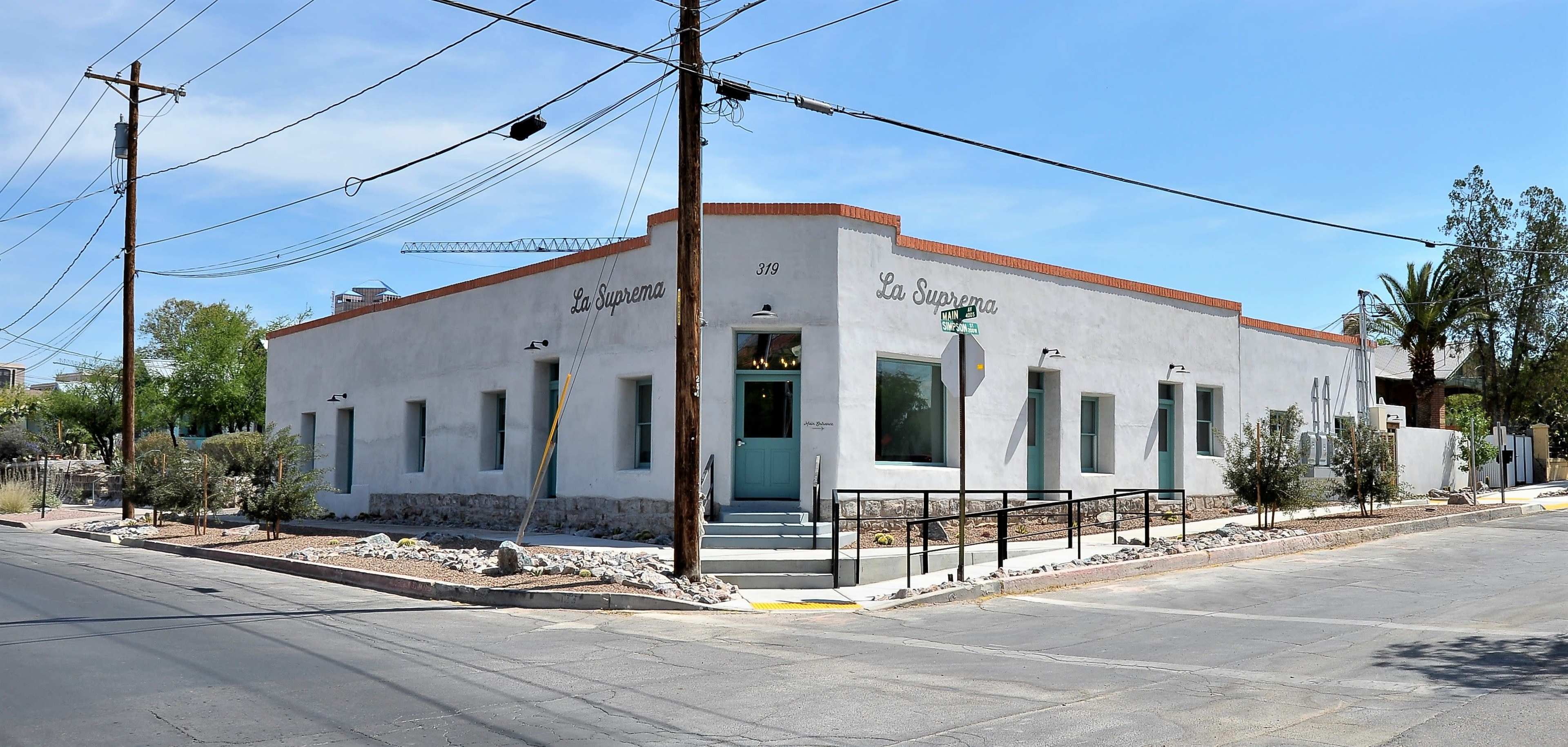 A single-story, white stucco building with a red-brick trim and large windows is located at the corner of a street, featuring a ramp for accessibility and surrounded by landscaped gravel and shrubs.