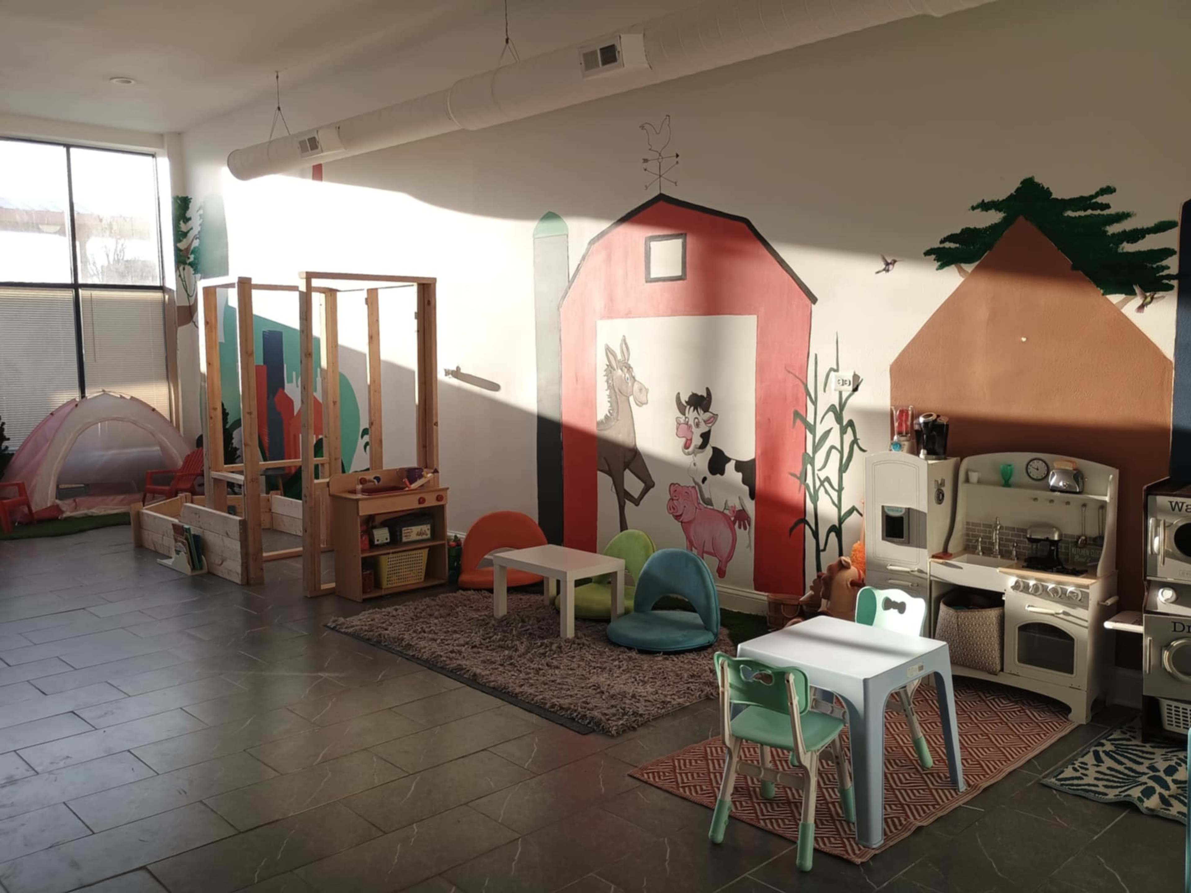 The image shows a bright playroom with wooden toys, a small kitchen set, and colorful seating arranged around a patterned rug, all against a mural featuring farm animals.