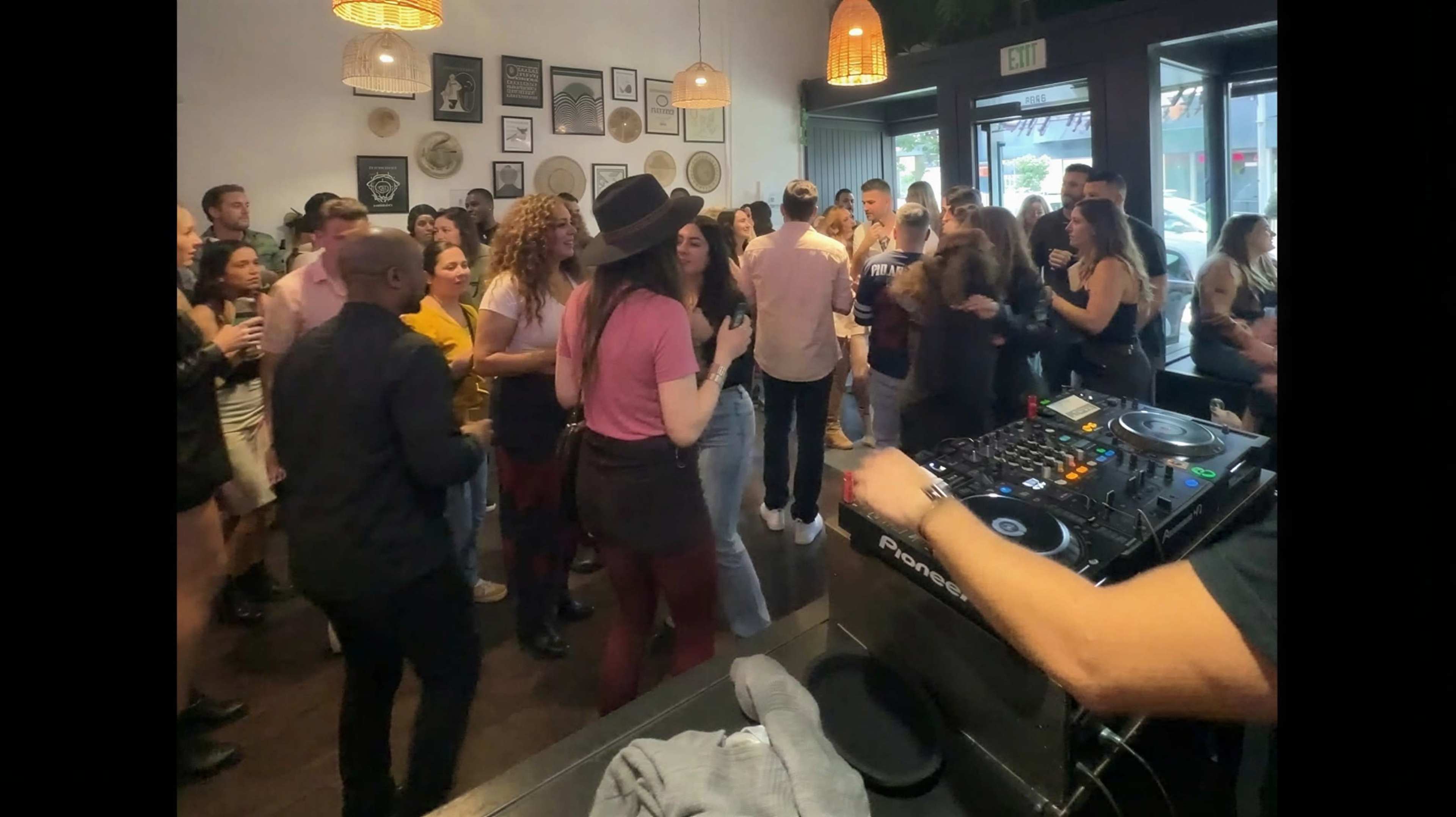 A crowd of people is dancing and socializing in a lively indoor venue with a DJ setup in the foreground.
