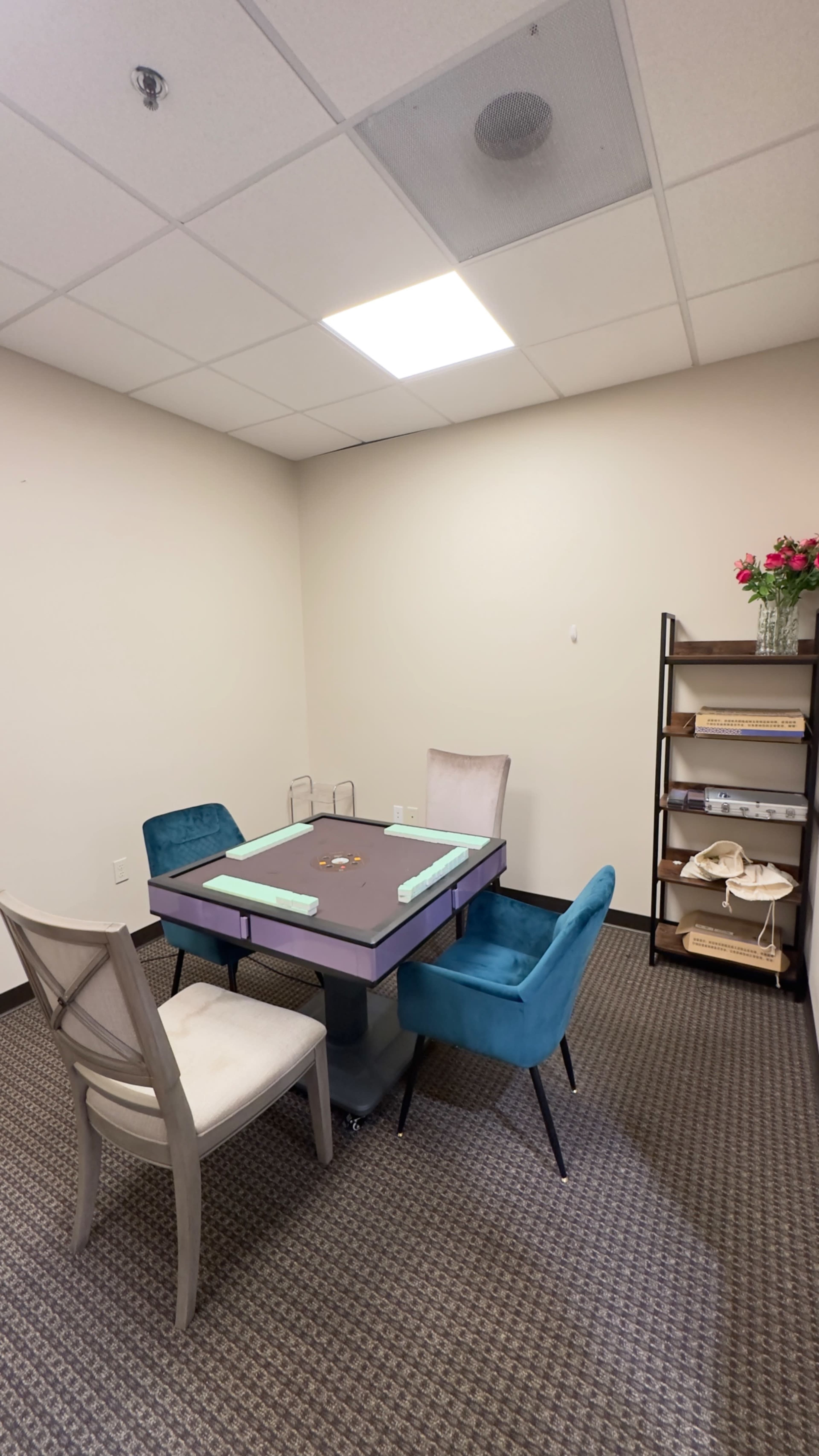 The image shows a small, empty meeting room with a table and four chairs, along with a shelving unit containing decorative items.