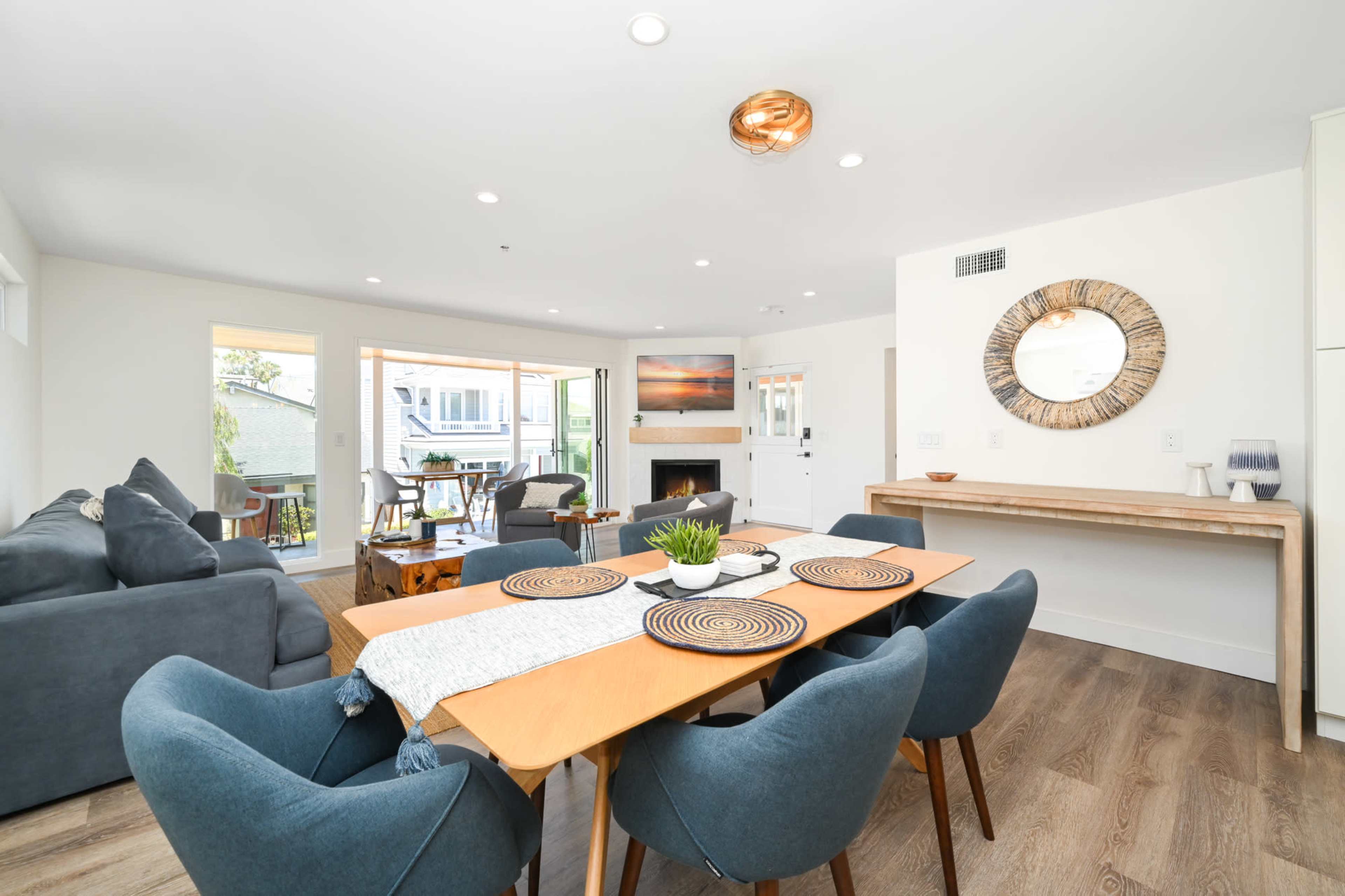 A modern living and dining area features a wooden table with blue chairs, a gray sofa, and large windows that open to a balcony.