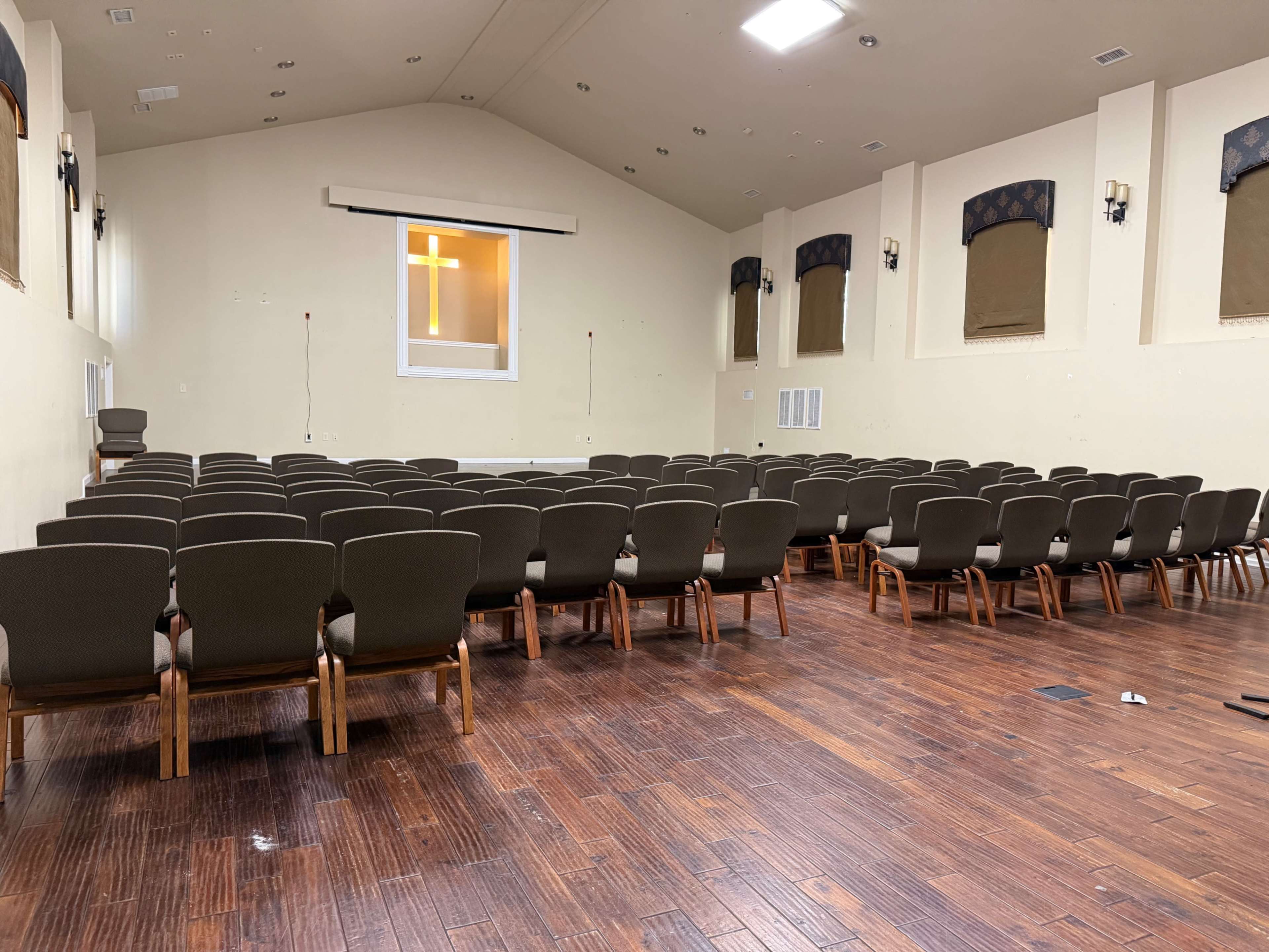 The image shows an empty auditorium with rows of chairs facing a front stage area, featuring a large window with a cross visible.
