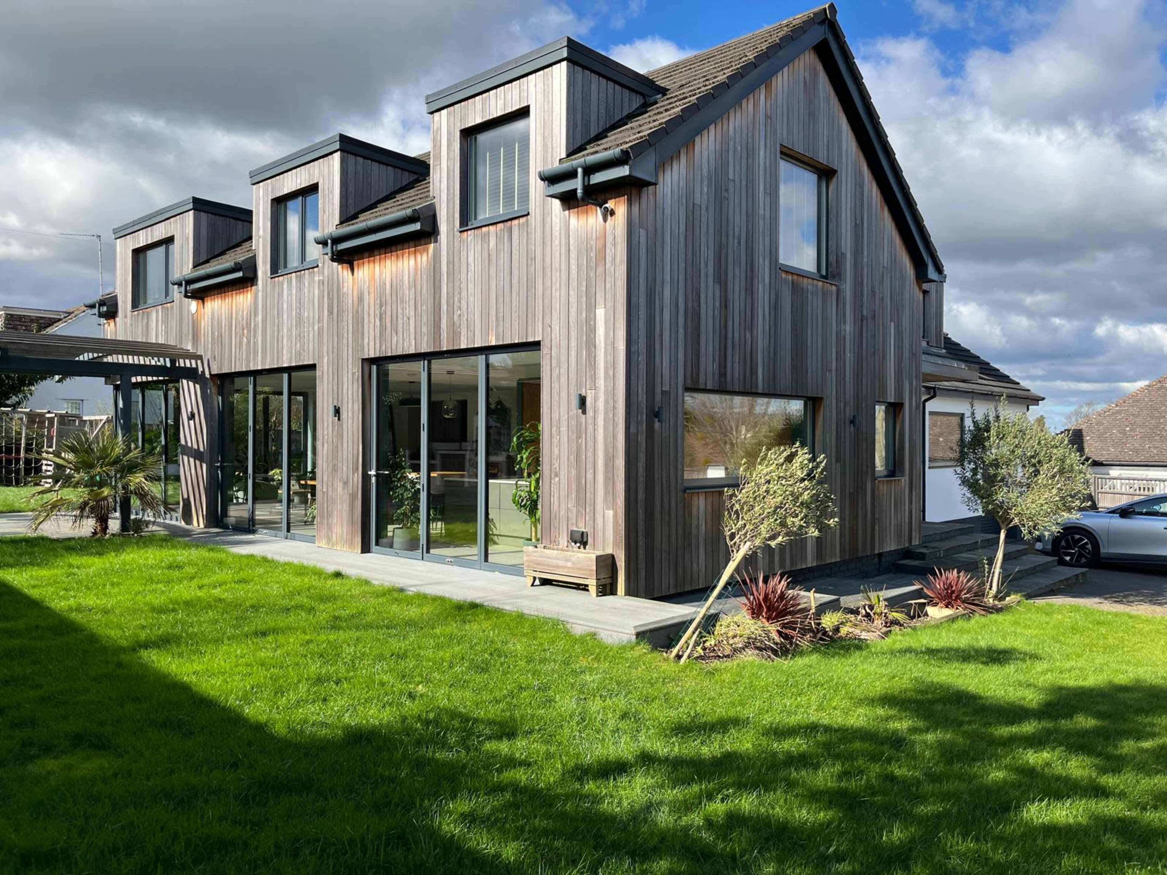 The image shows a modern wooden-clad house with large glass windows, set on a green lawn.