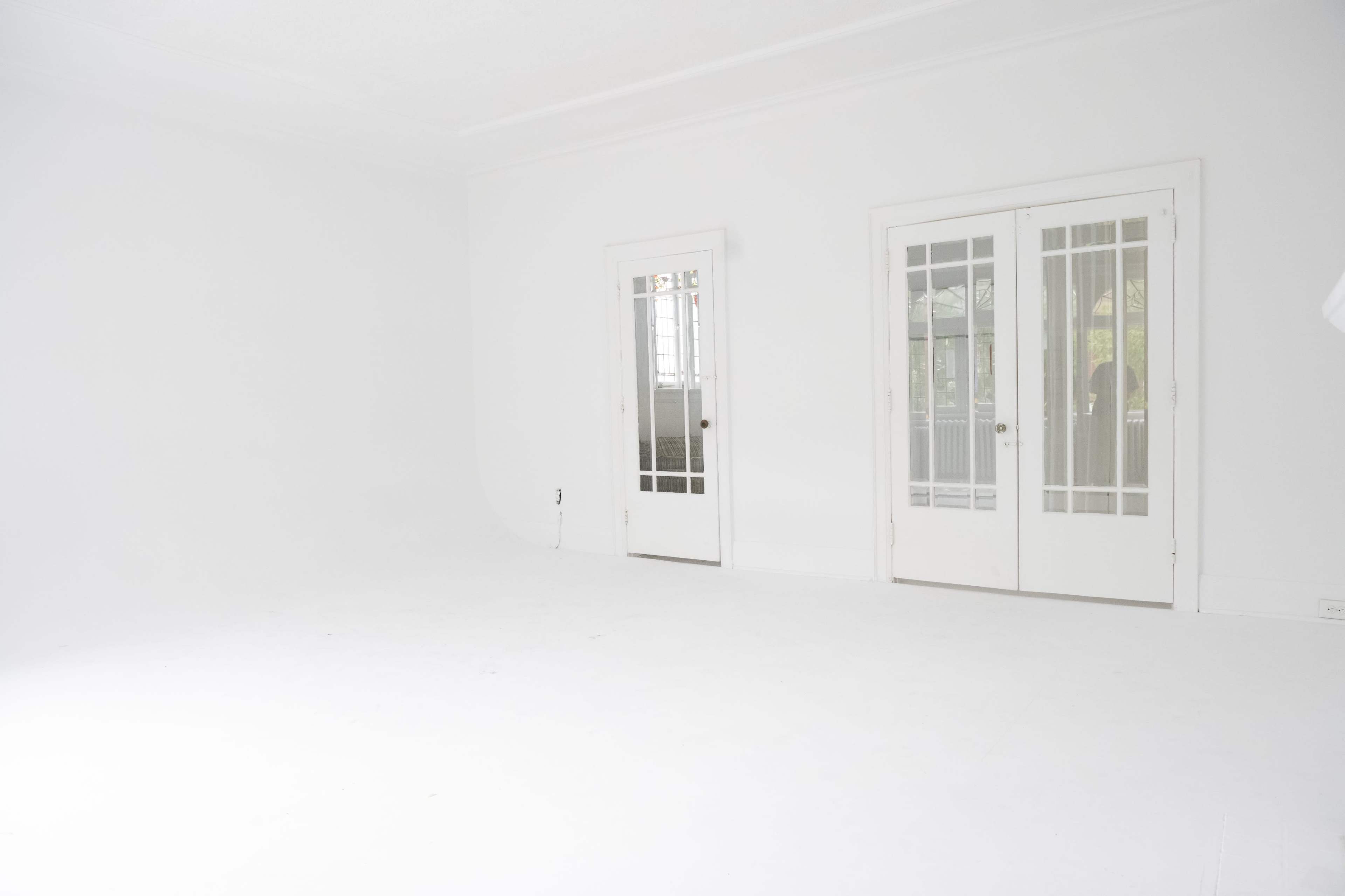 A spacious, empty room with white walls and flooring, featuring two glass-paneled doors leading to an exterior area.