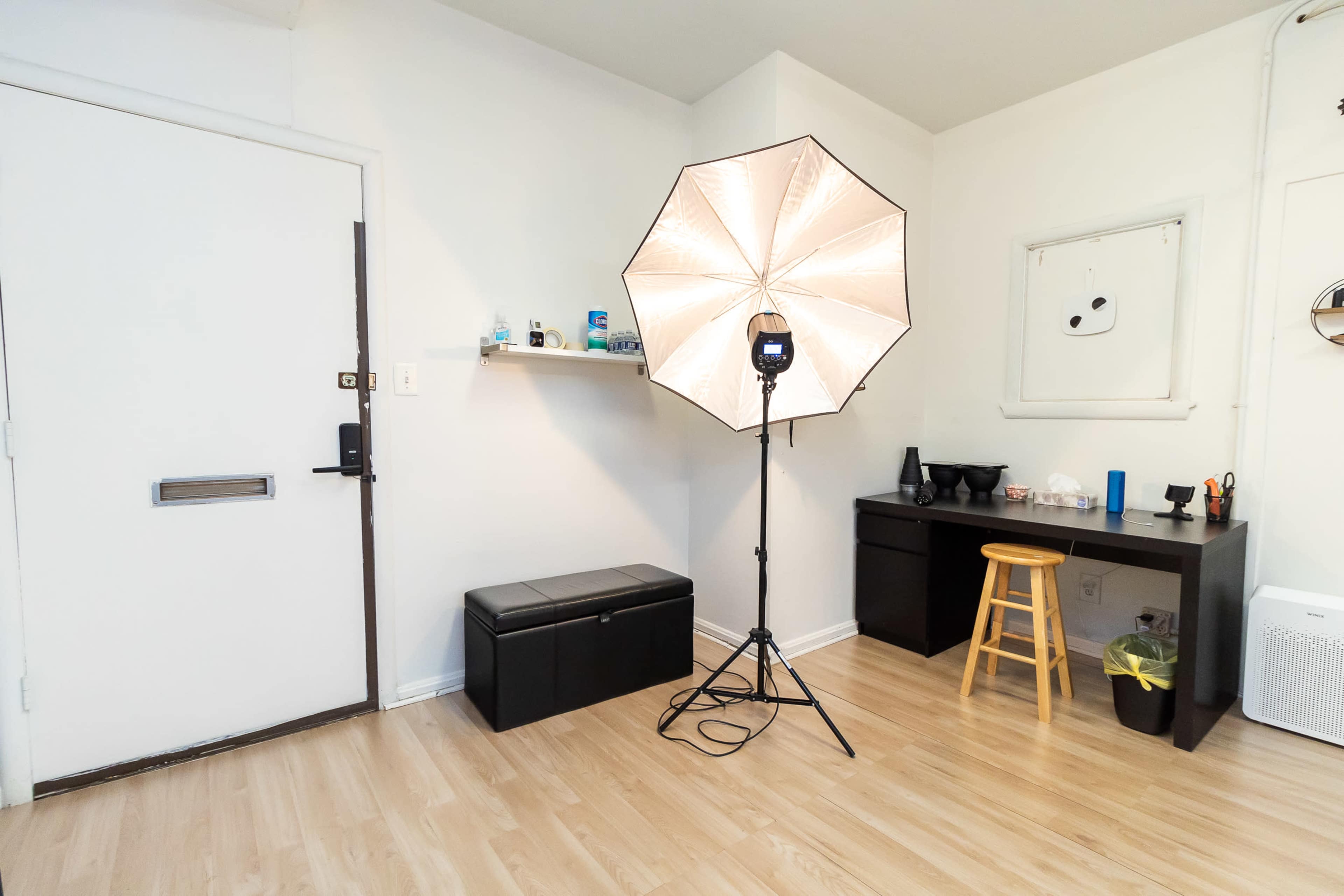 The image shows a well-lit room featuring a large softbox light on a stand, a black storage bench by the door, and a dark desk with a few items neatly arranged on it.