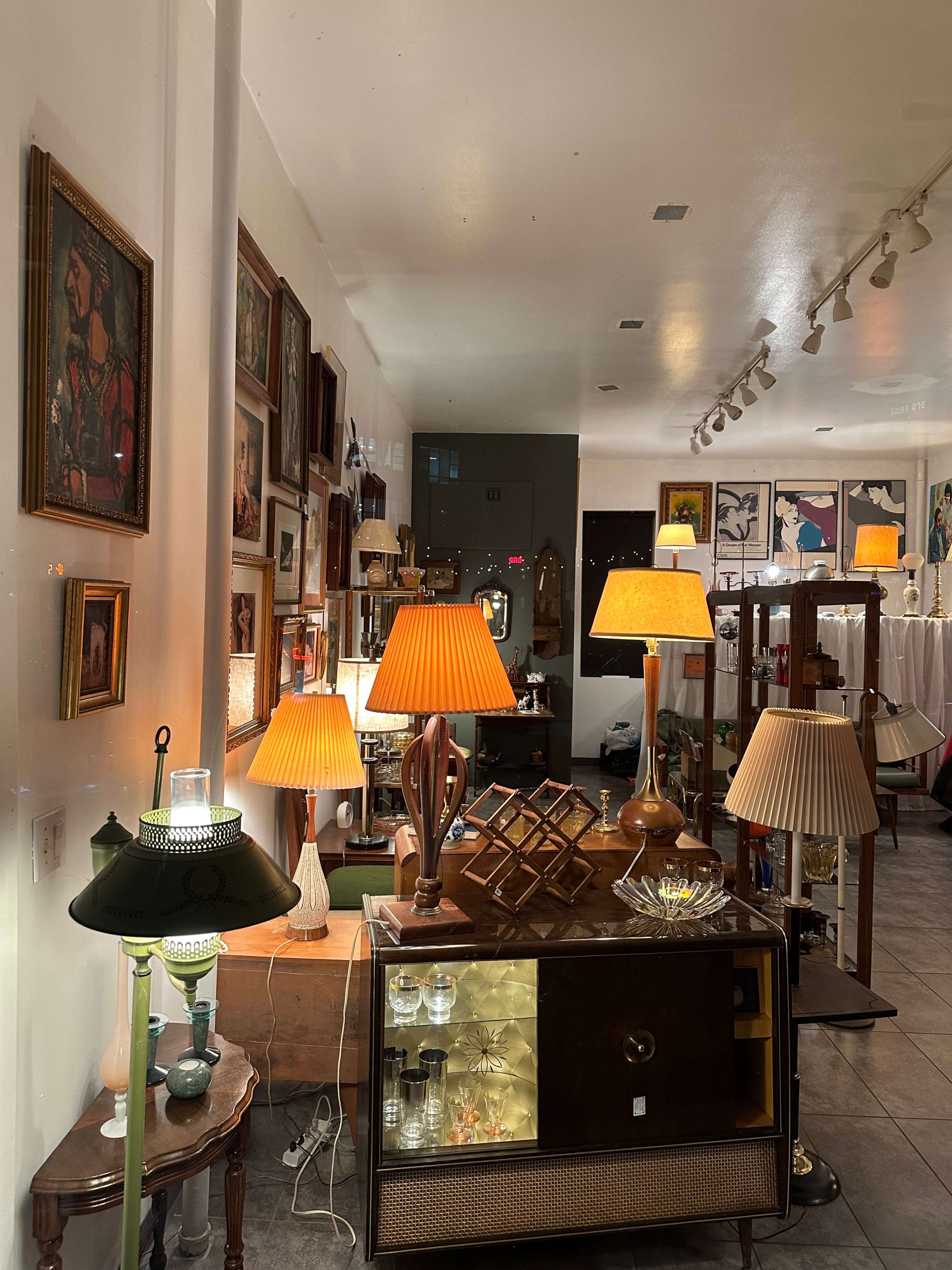 The image shows a well-lit interior of an antique shop featuring various lamps and framed artwork on the walls.