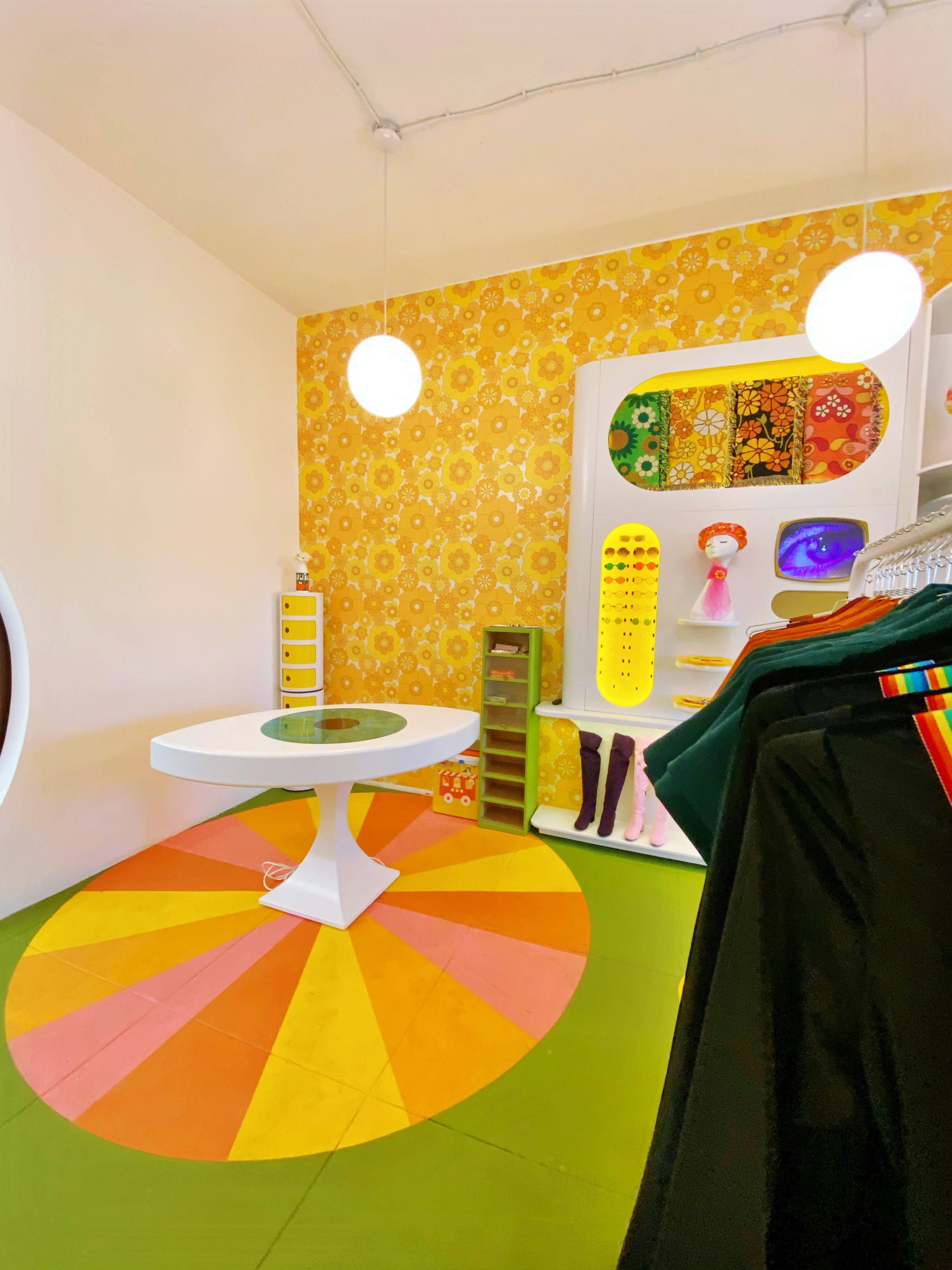The image shows a colorful boutique with a circular table in the center, a patterned wall, and displays of clothing and accessories.