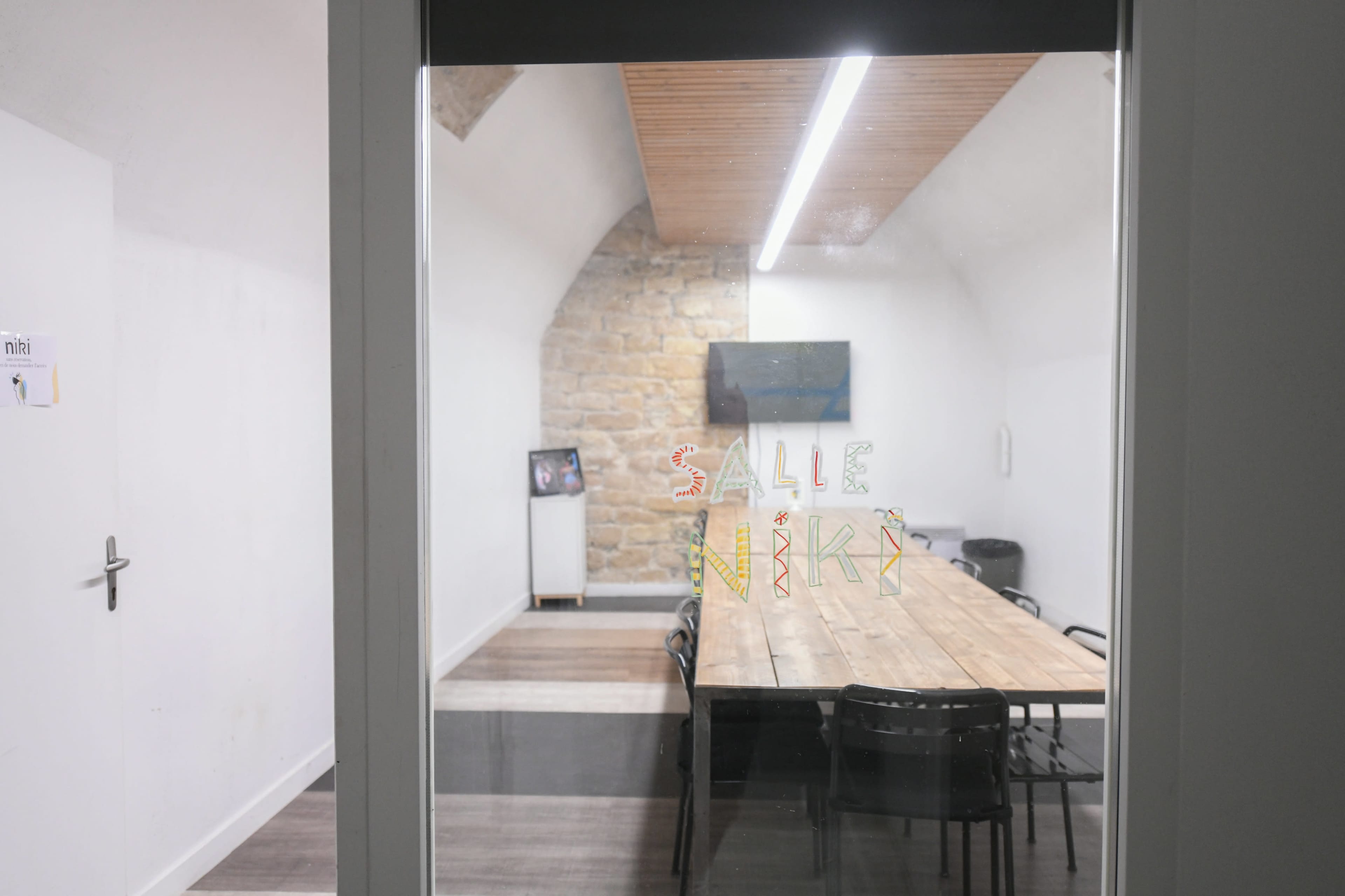 A meeting room with a long wooden table and black chairs, viewed through a glass door with the name "Salle Niki" on it.