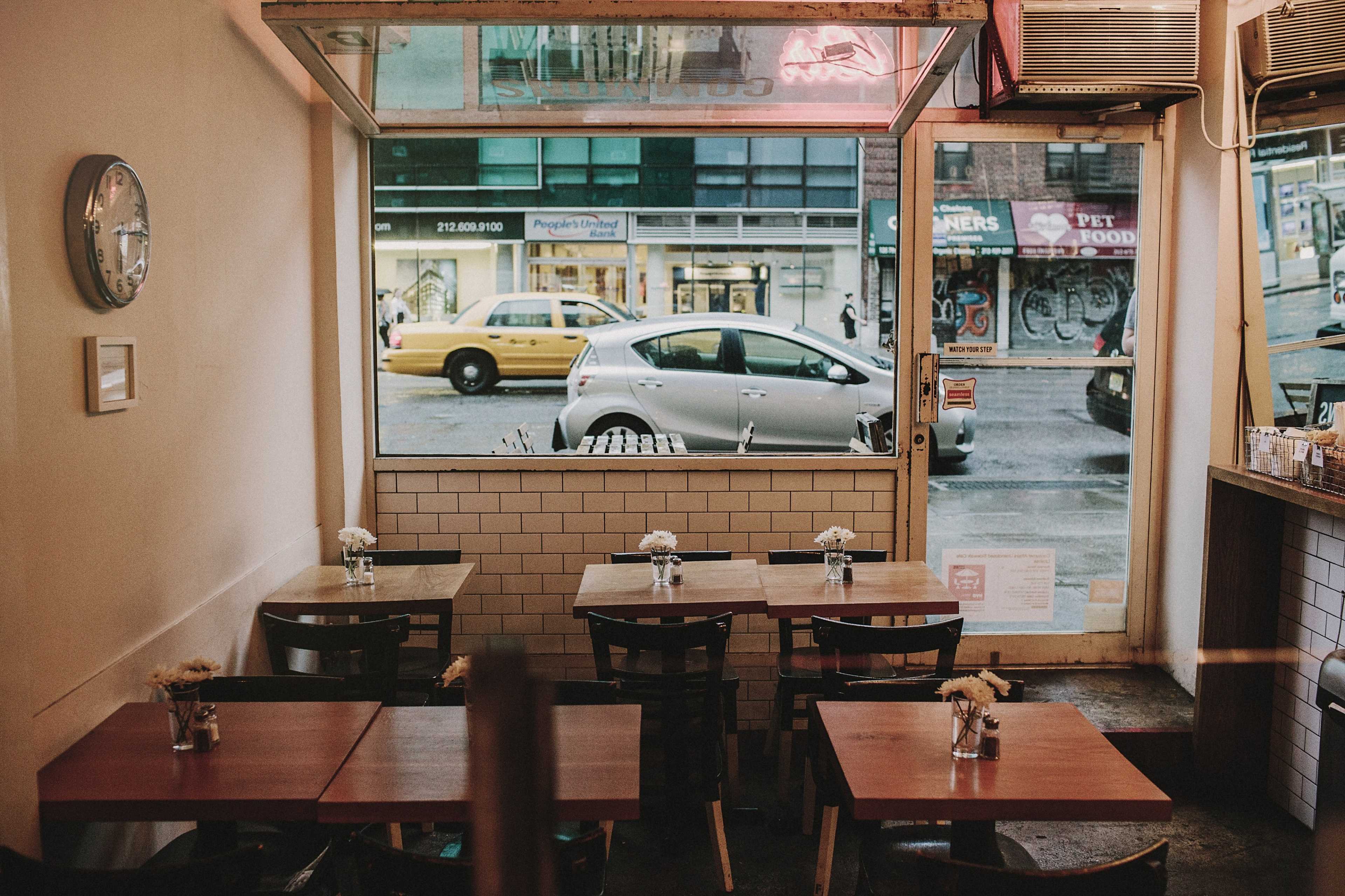 A small café with empty wooden tables and a view of a street with a parked car and a yellow taxi outside.