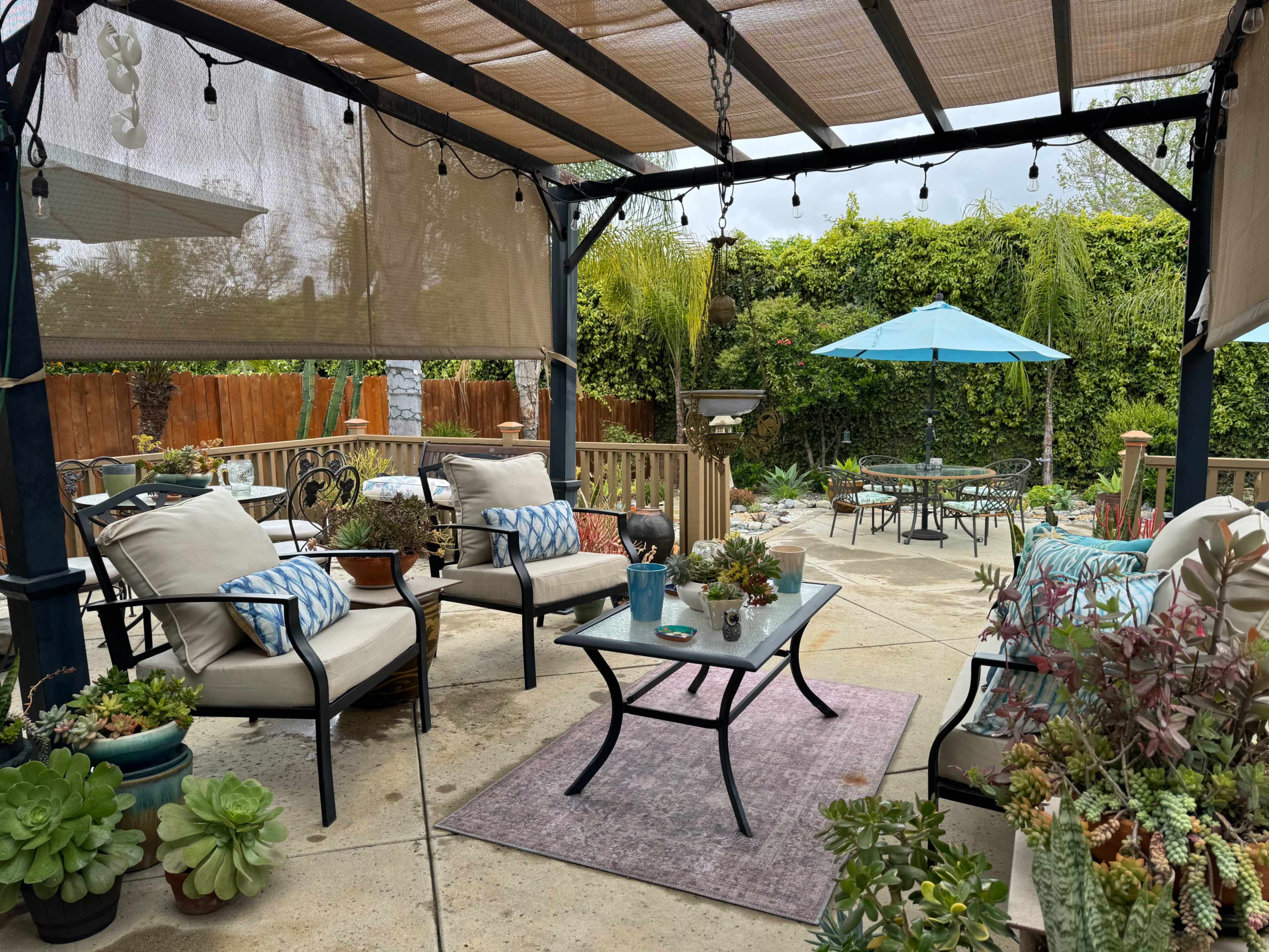 A covered patio features seating arrangements and potted plants, with a table in the center and an umbrella over a dining area in the background.