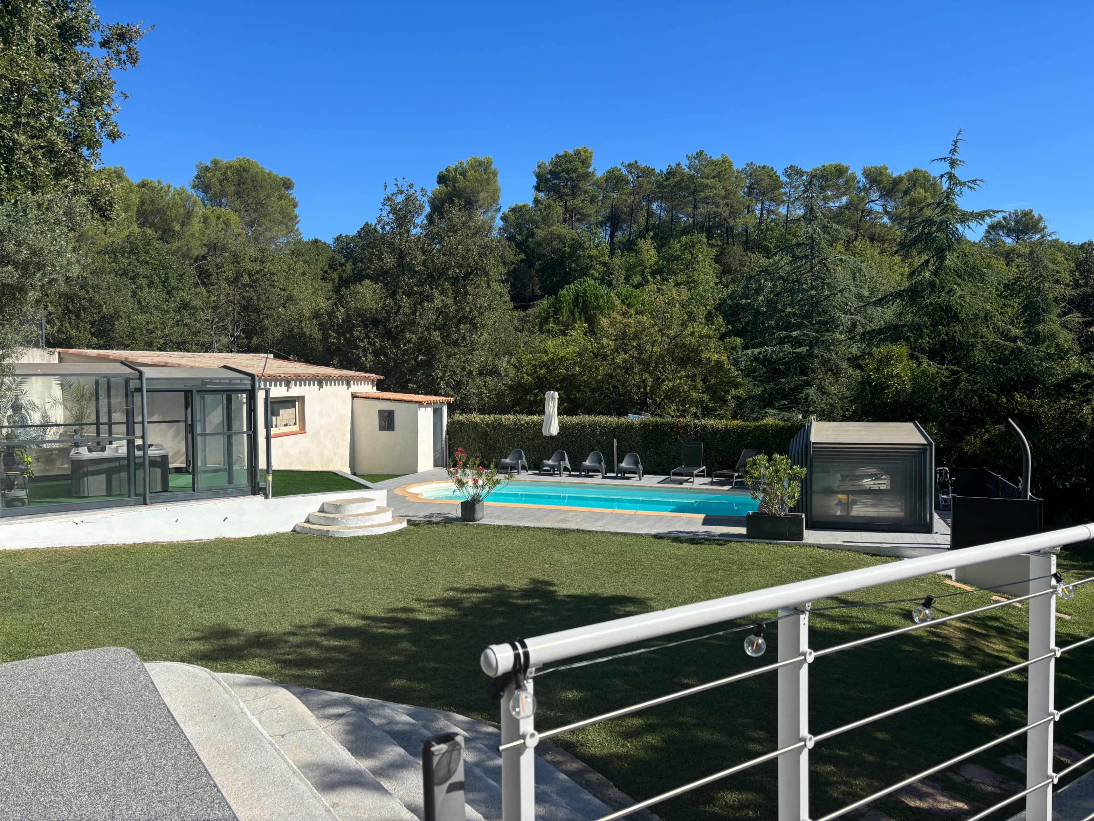 The image shows a modern backyard featuring a swimming pool, lounge chairs, a glass-enclosed area, and a landscaped garden surrounded by trees.