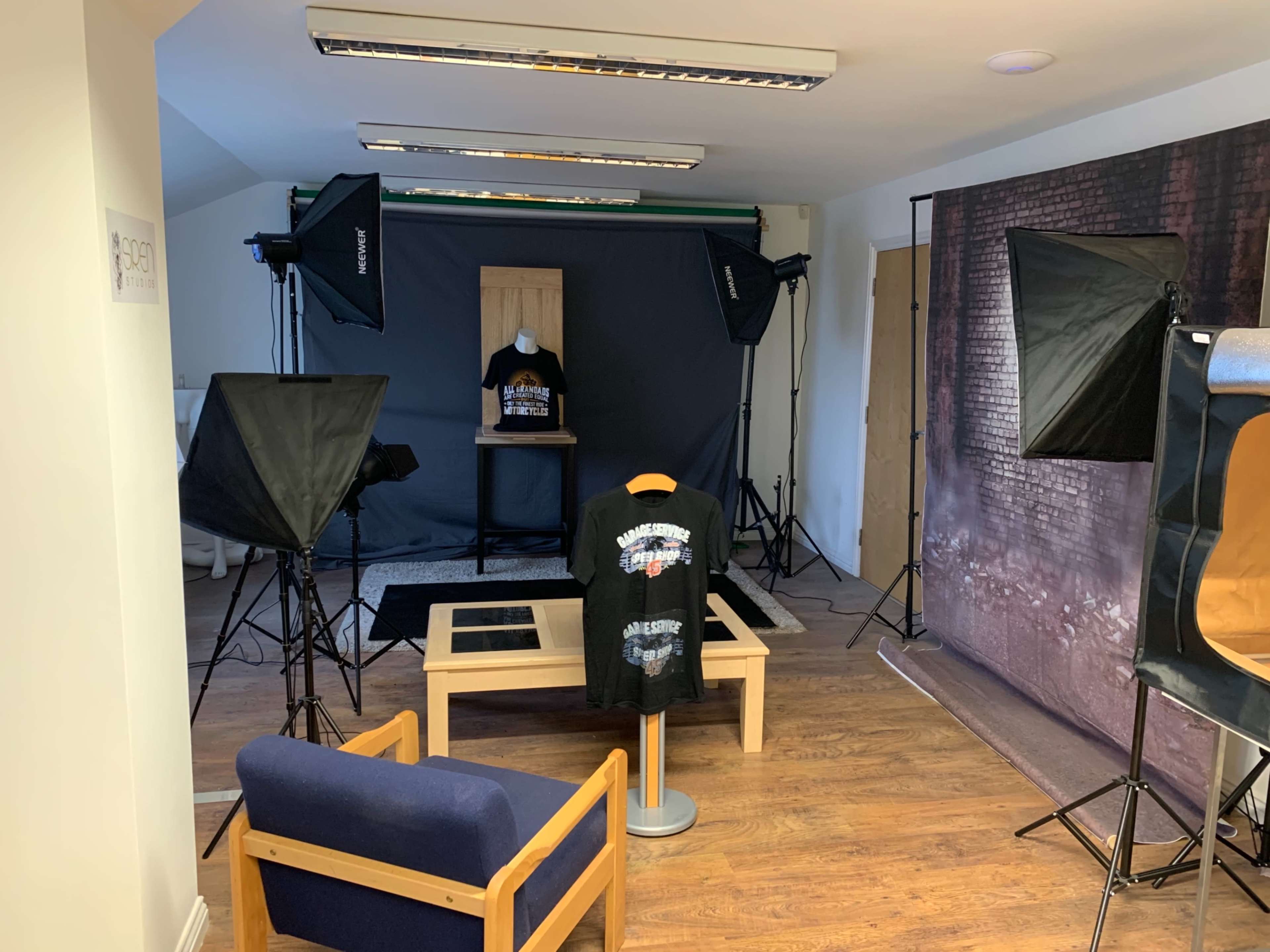 The image shows a photography studio setup with various lighting equipment, a backdrop, and a display of a T-shirt on a mannequin and a table.