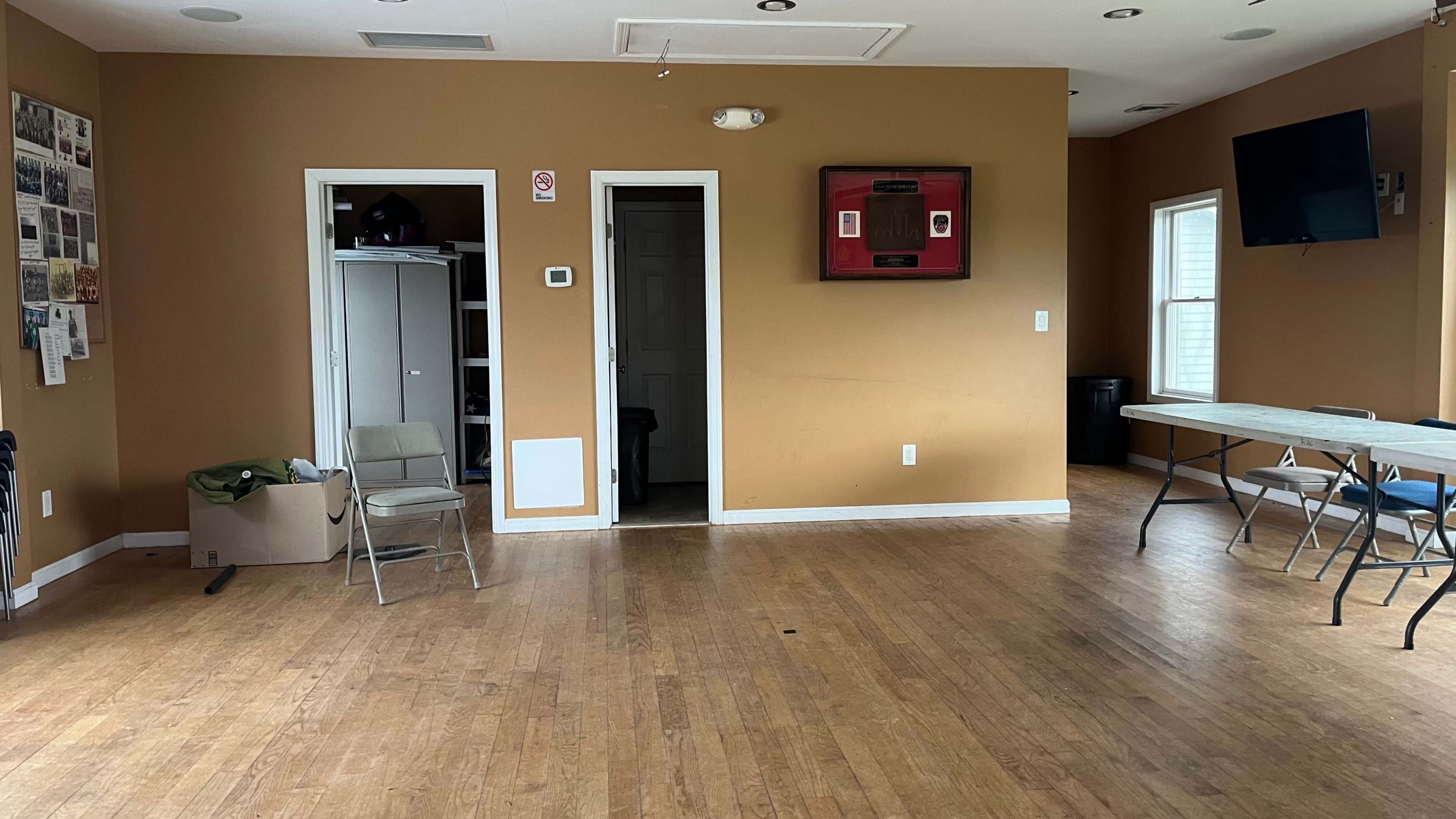 The image shows an empty room with brown walls, a door leading to another room, a wall-mounted television, and a few chairs and tables arranged inside.