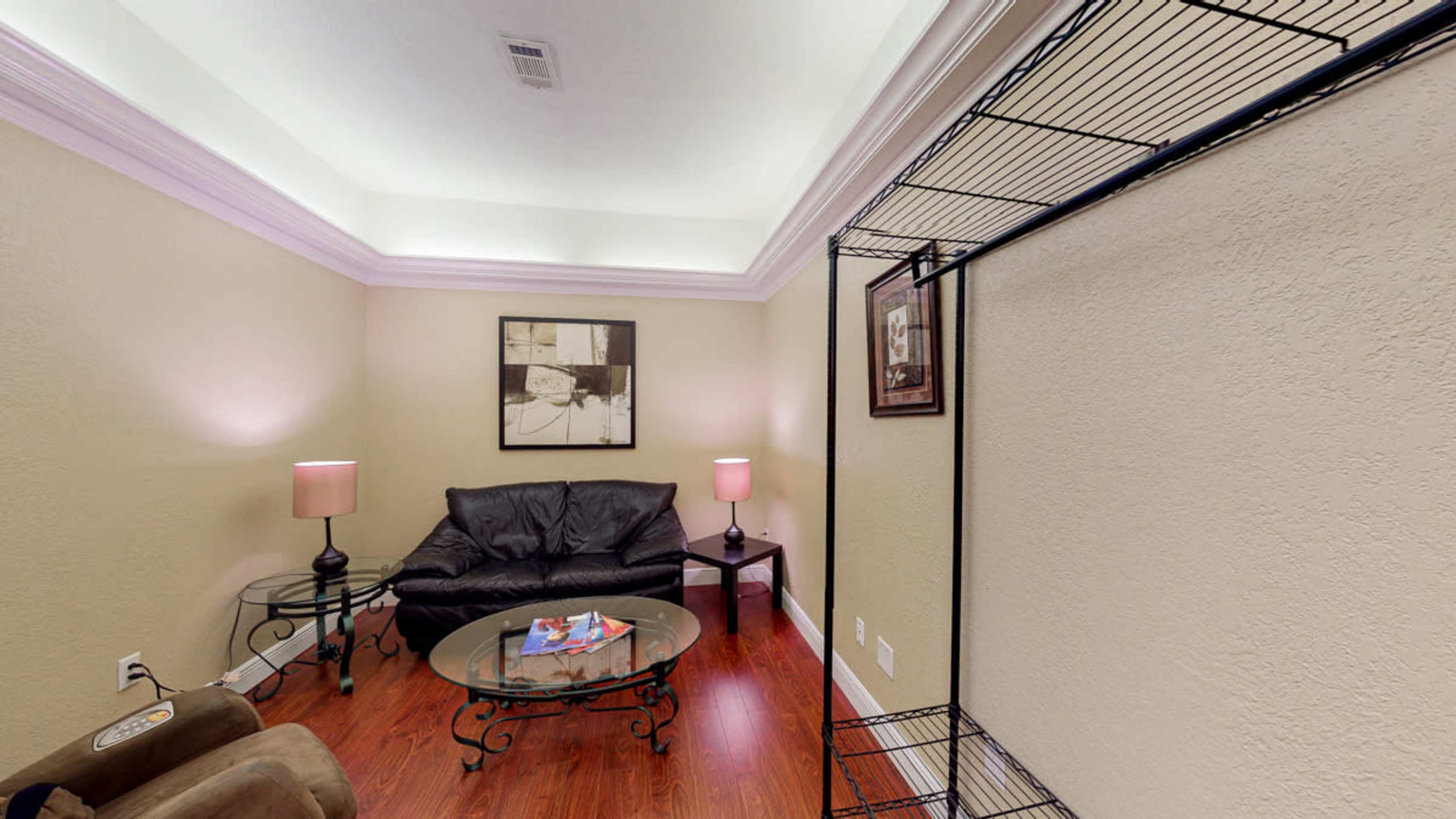 The image shows a small, neatly arranged room with a black leather couch, a glass coffee table, and two lamps providing soft lighting.