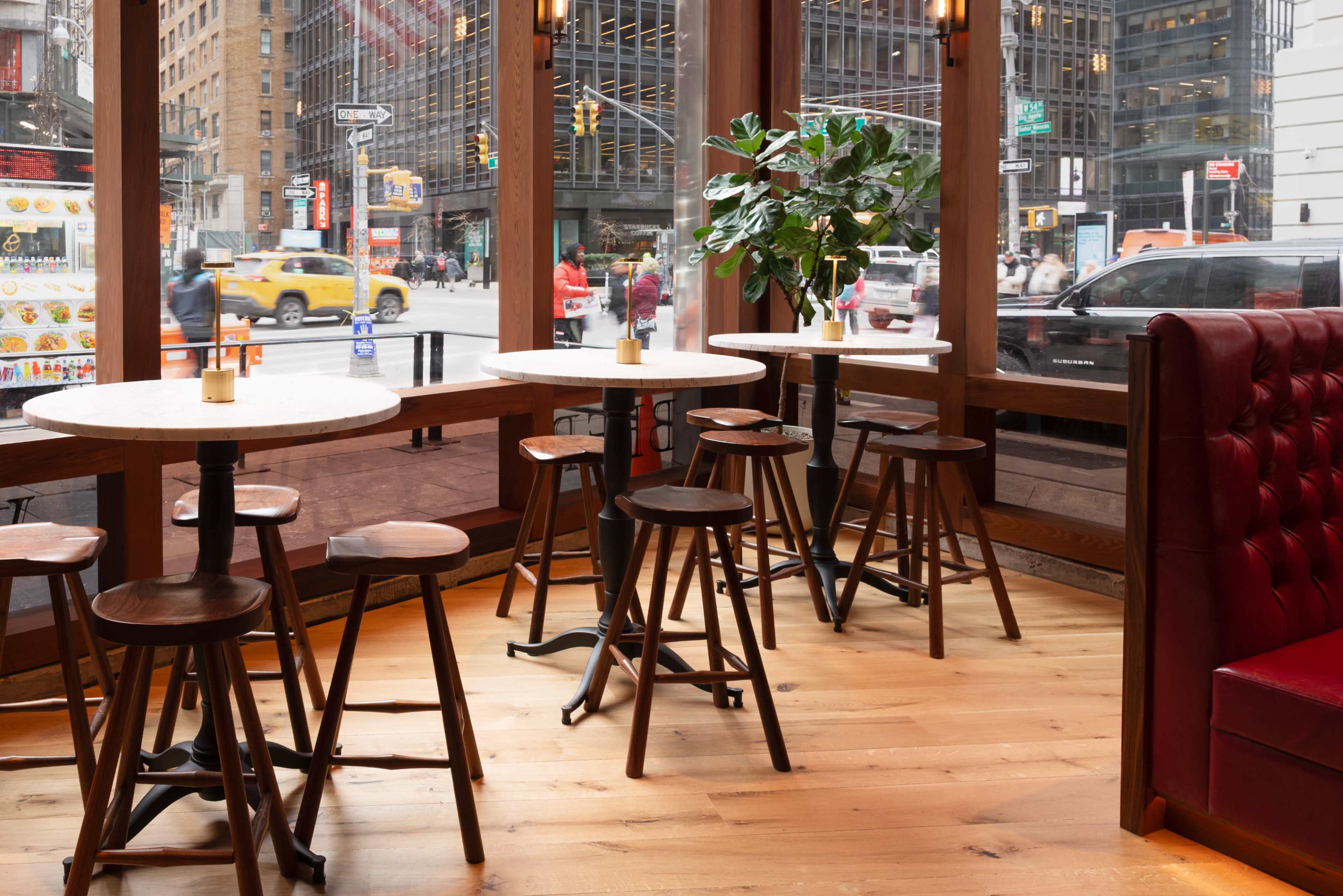 The image shows a restaurant interior featuring a few round tables with wooden stools, large windows overlooking a busy city street, and a potted plant in the corner.