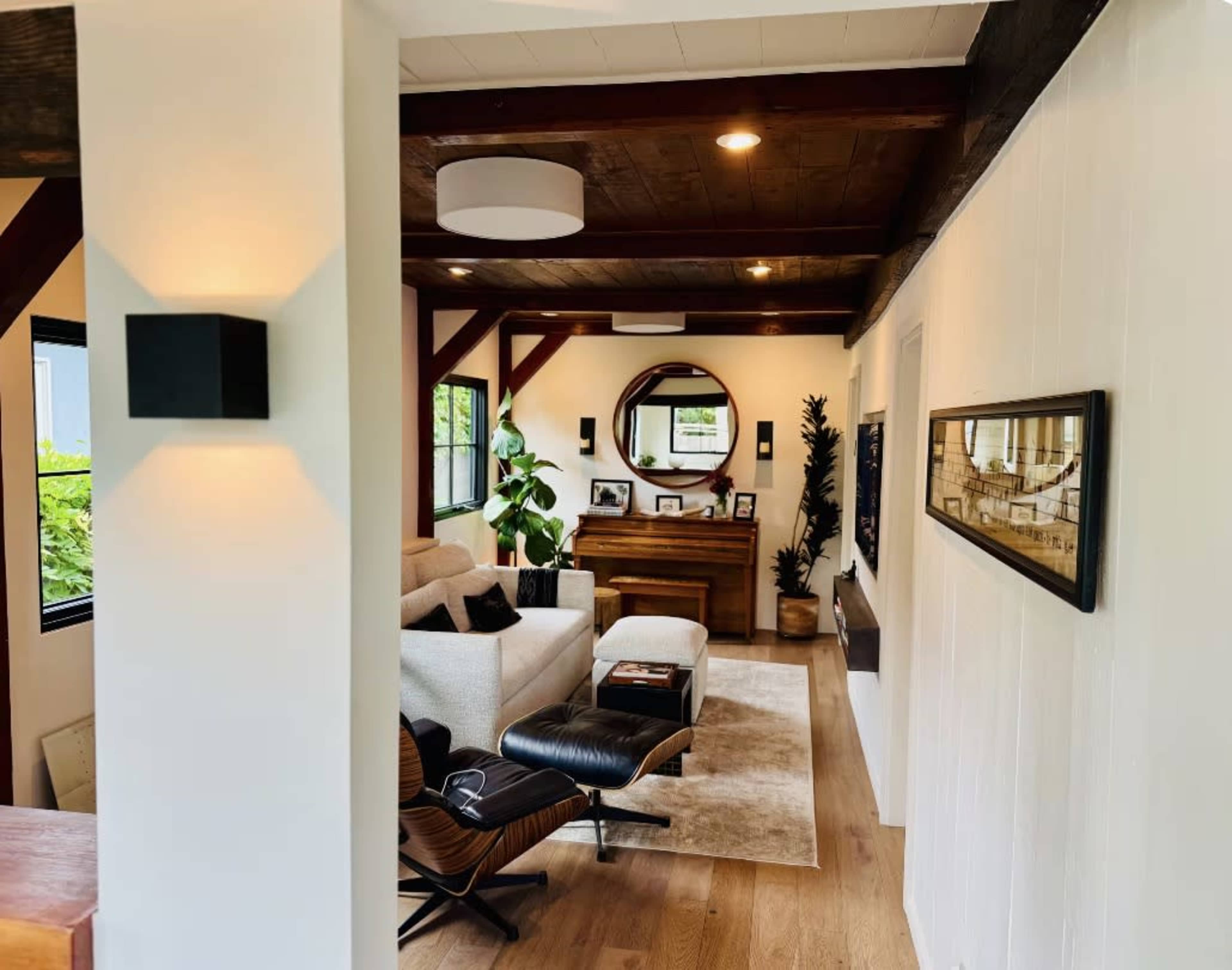 The image shows a modern living room with a light-colored sofa, a wooden piano, and a circular mirror, complemented by plants and contemporary decor.