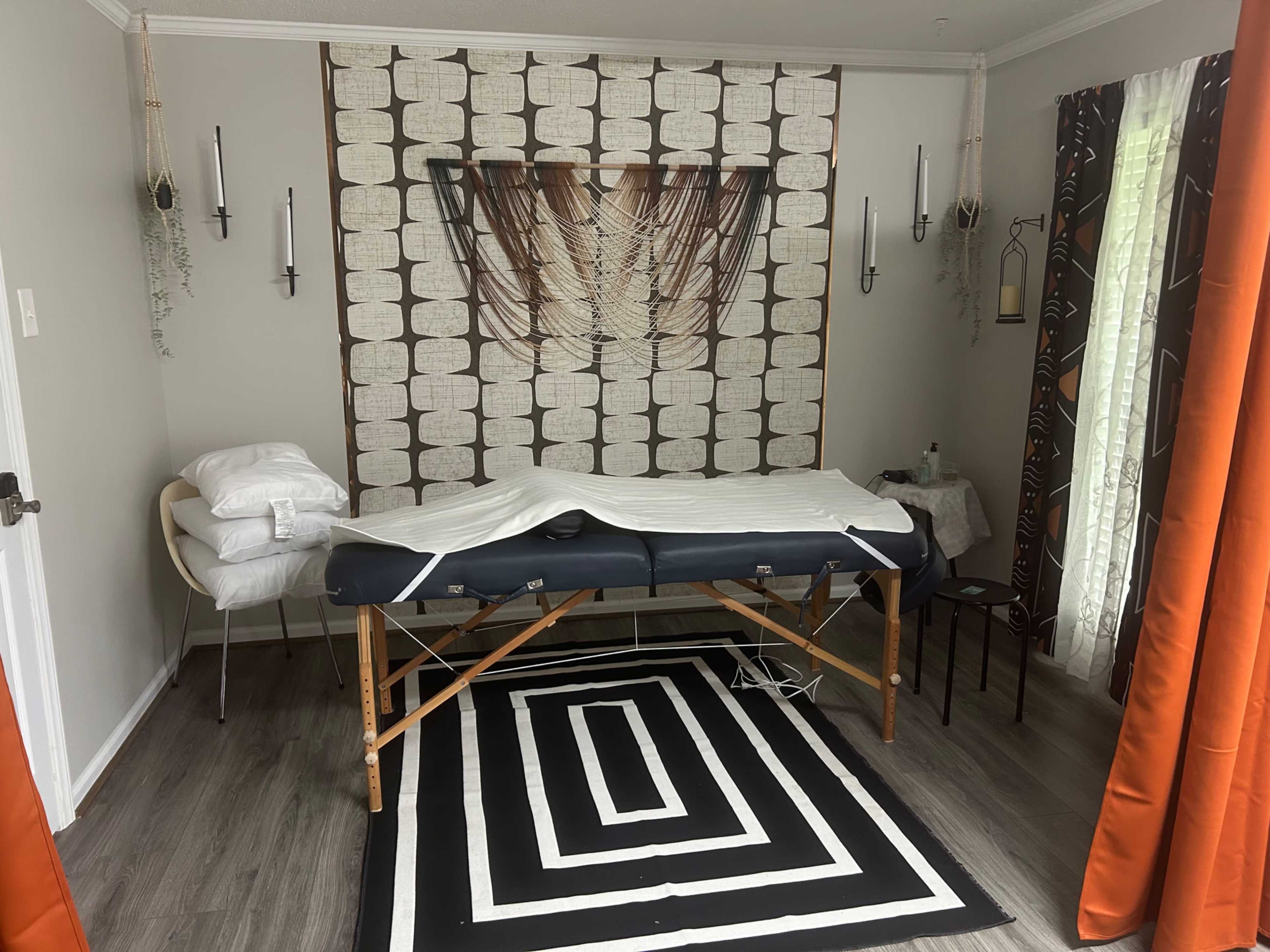 The image shows a massage therapy room with a therapy table, a chair, and decorative wall hangings.