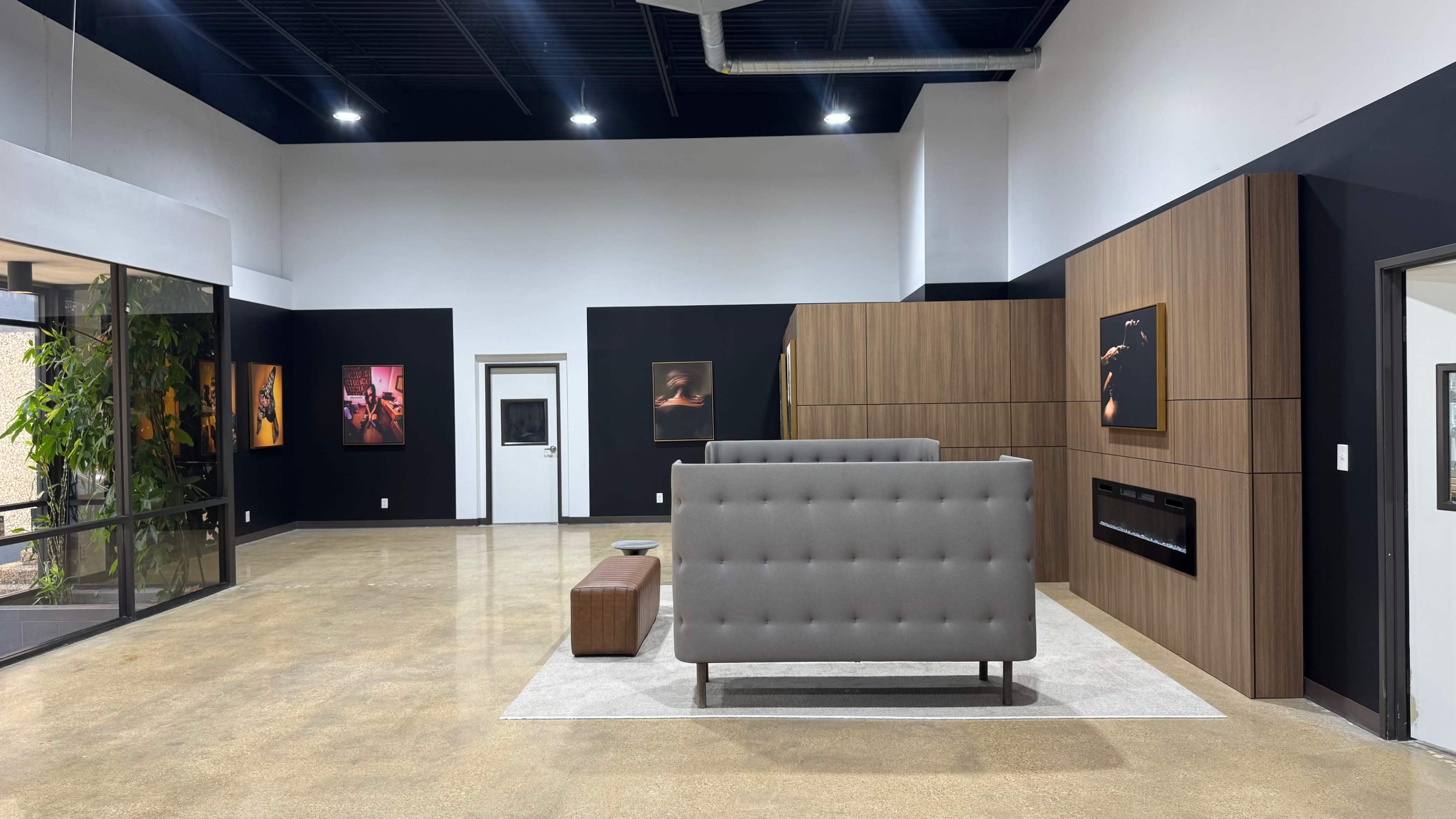 The image shows a modern interior space featuring a gray fabric sofa, a brown leather ottoman, and artwork displayed on black walls.
