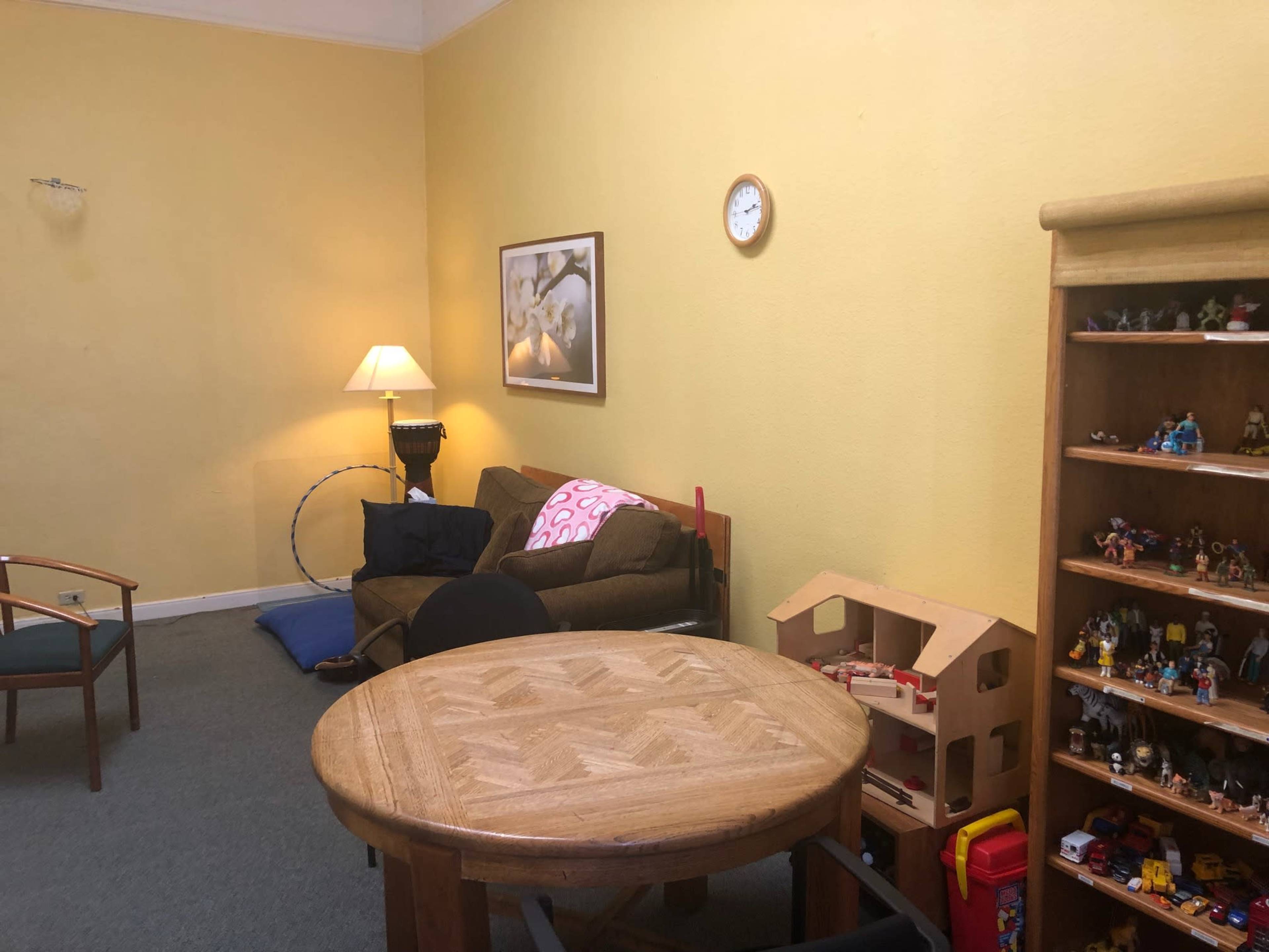 The room features a yellow wall, a brown sofa with a pink pillow, a wooden table, and a shelf filled with toys.