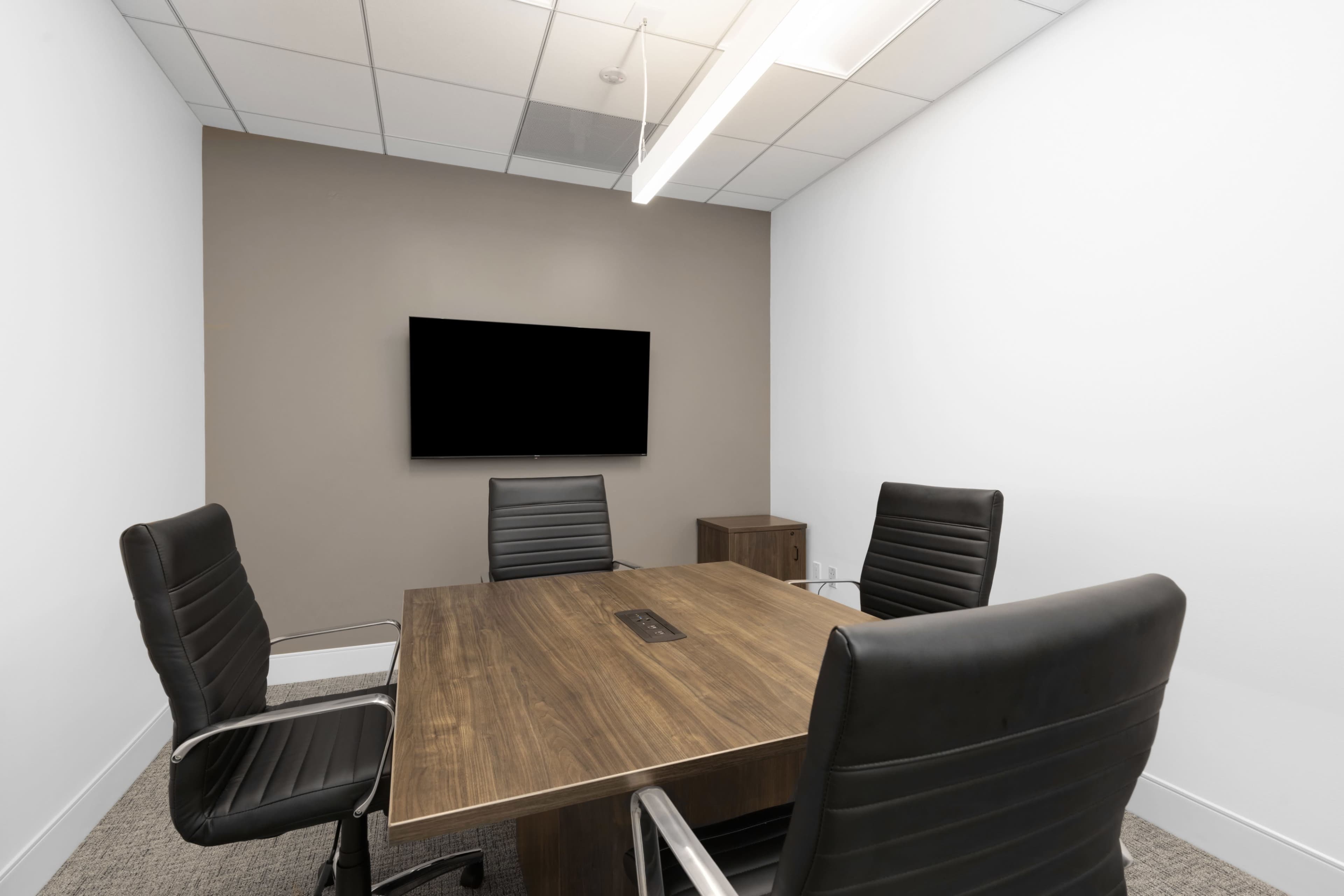A modern conference room features a wooden table surrounded by four black leather chairs and a wall-mounted television.