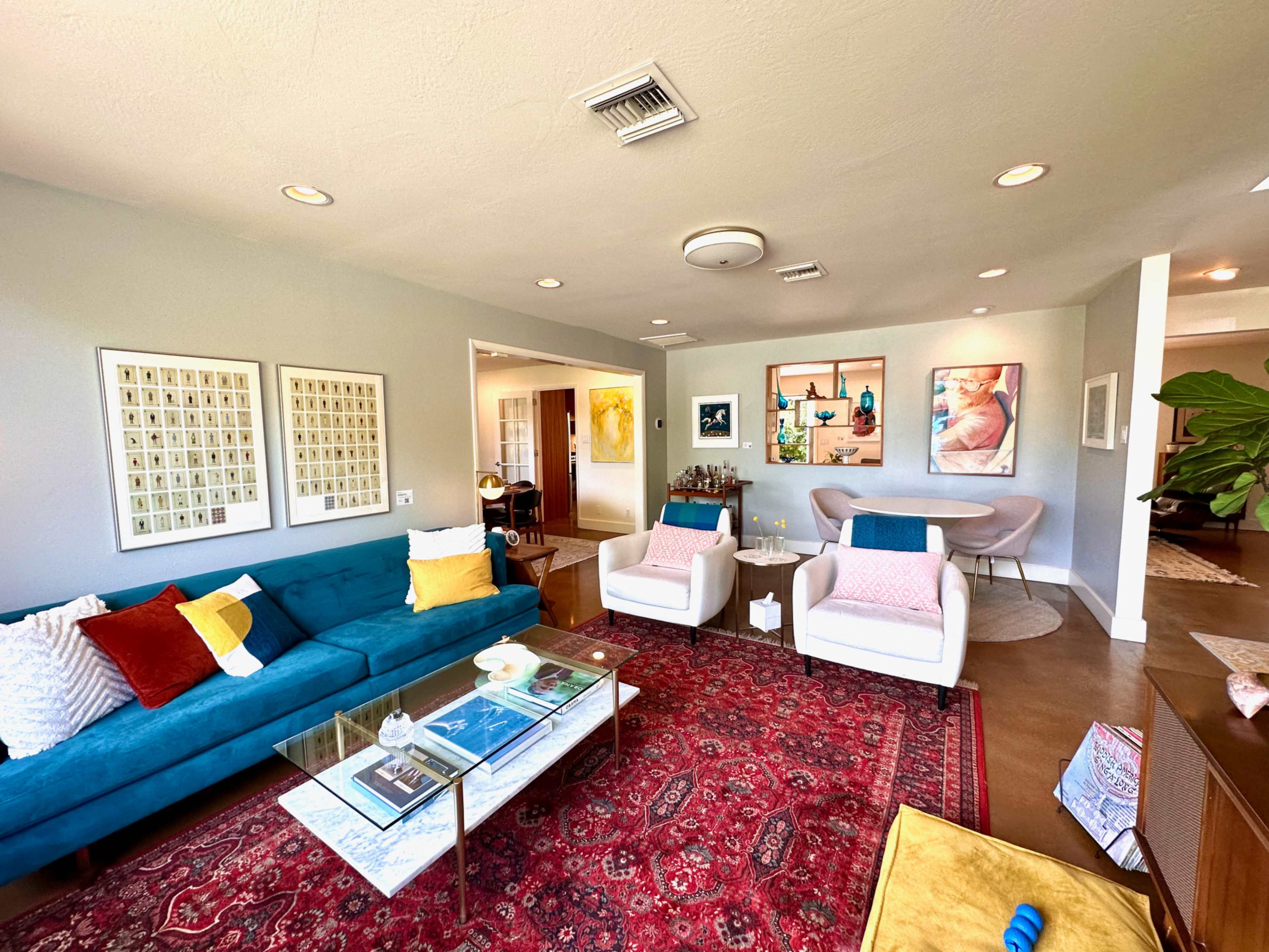 The living room features a blue sofa, two white armchairs, a glass coffee table, and a large area rug, with artwork and decorative items on the walls and surfaces.