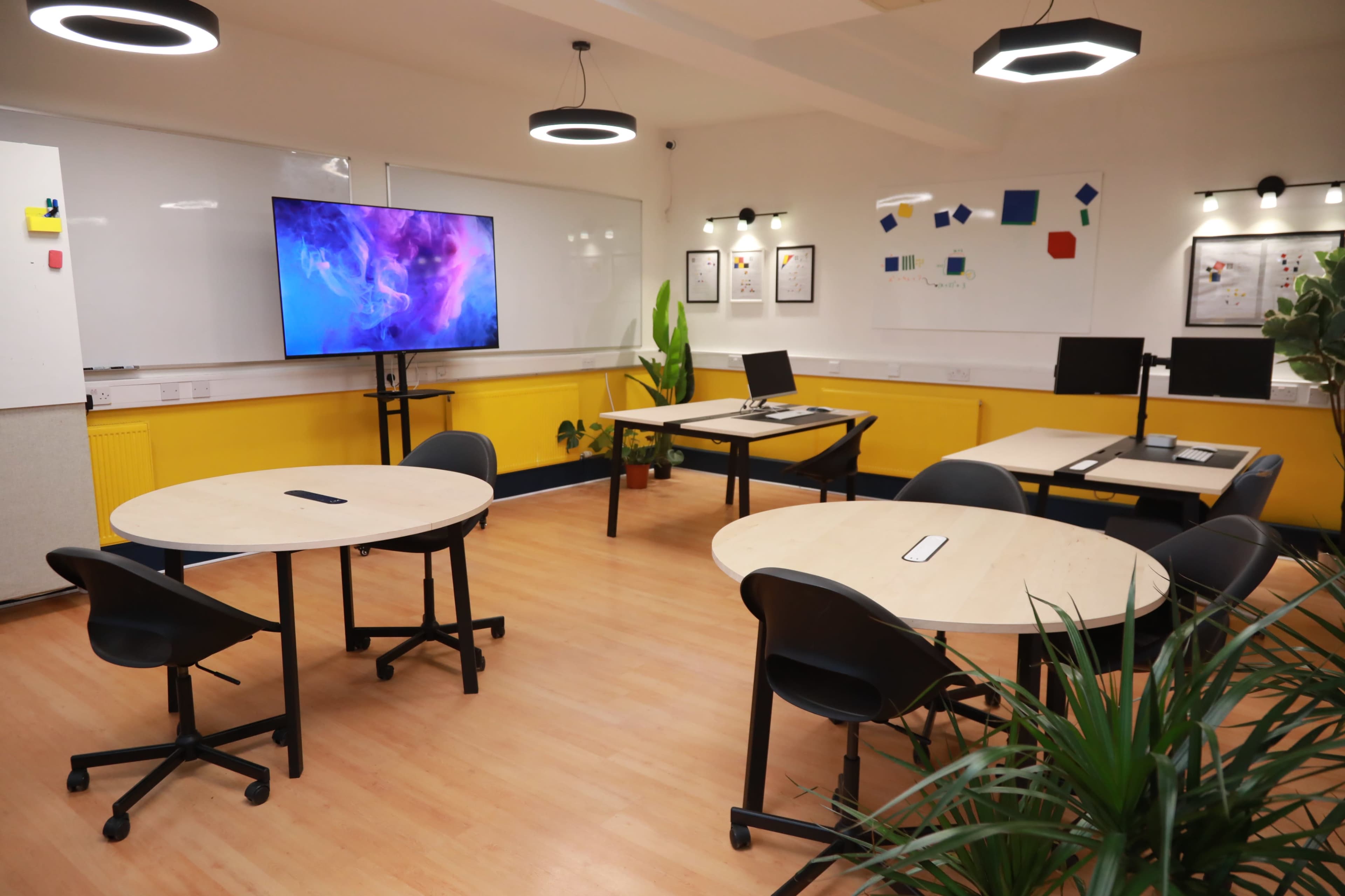 The image shows a modern classroom or meeting space with several round tables, chairs, a large screen, and colorful wall decorations.