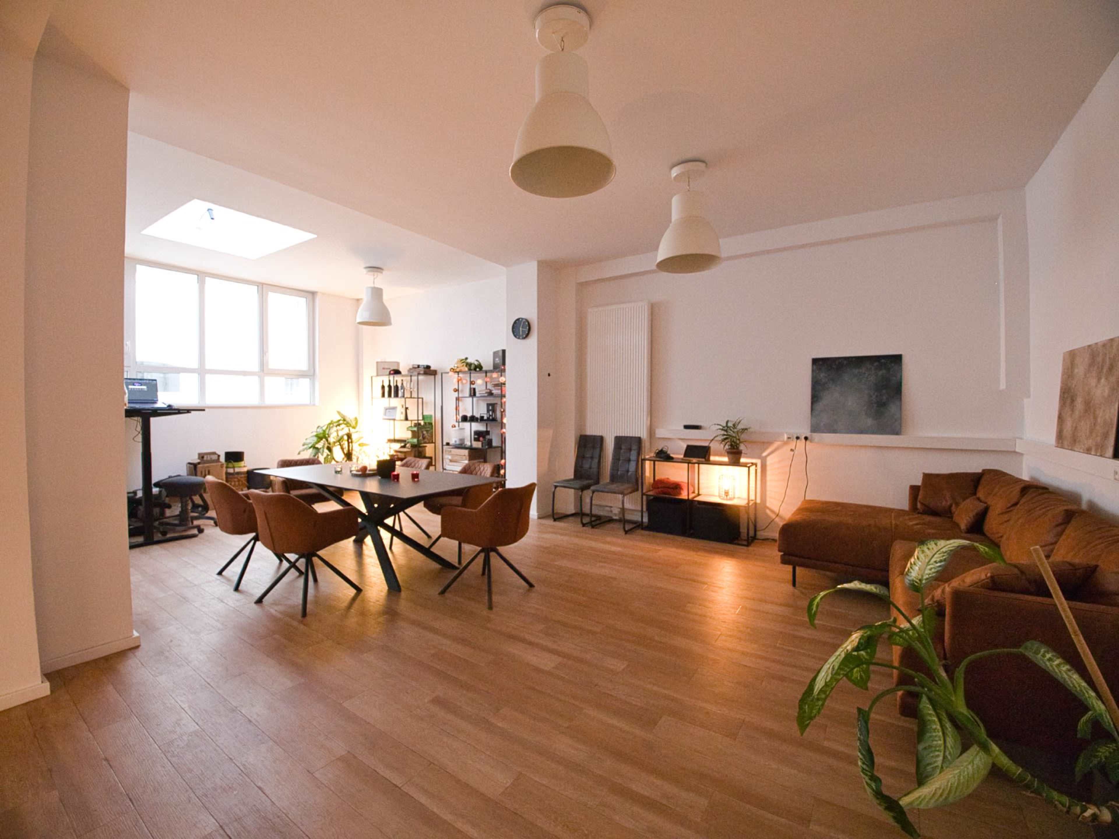 The image depicts a modern, open-concept office space with a dining table at the center, surrounded by several chairs, soft lighting, and a cozy seating area with a brown couch.