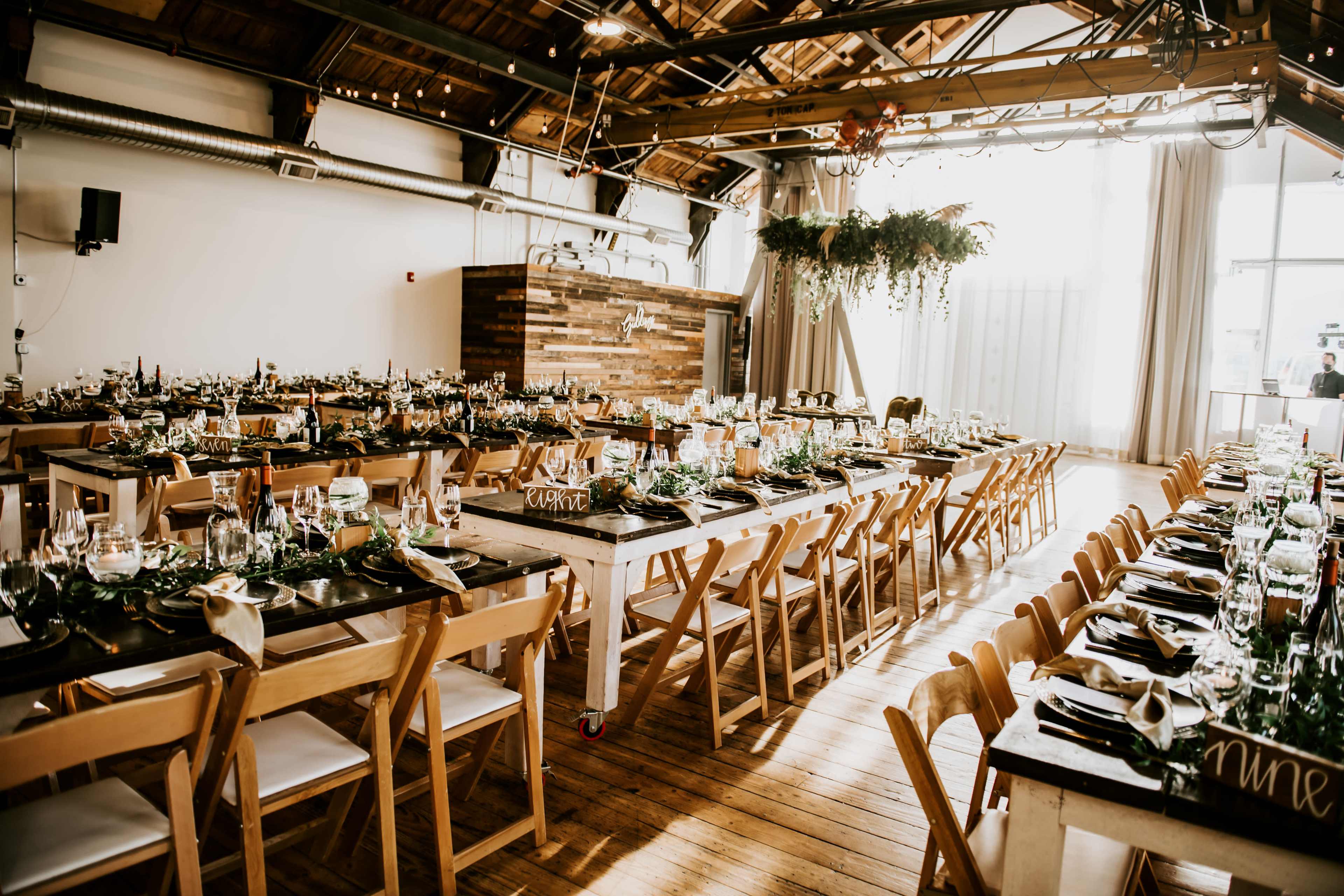 A large dining area featuring long wooden tables set with glassware, plates, and decorative greenery, under a high ceiling with wooden beams.