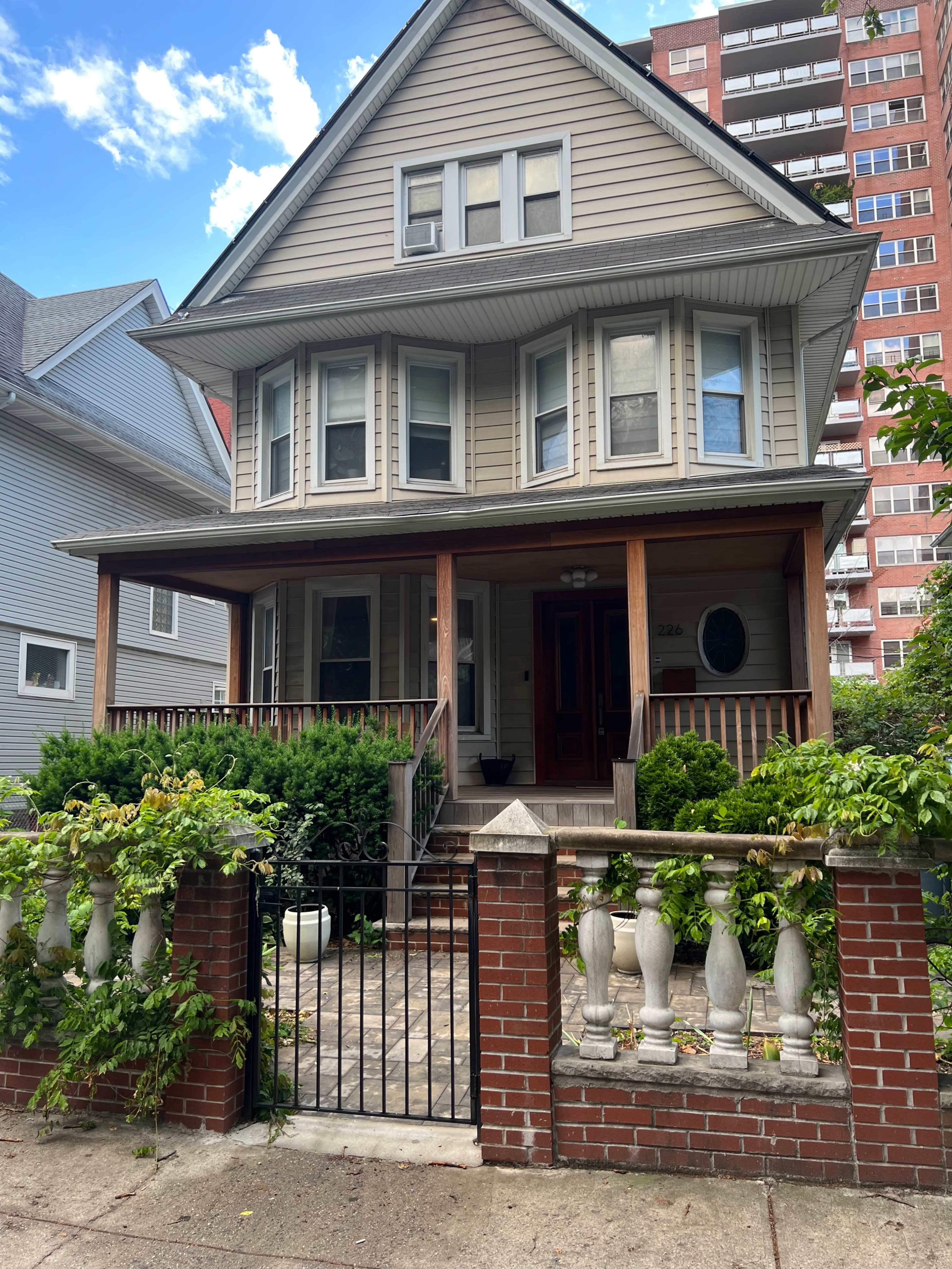 A three-story house with a front porch and decorative railing is situated behind a brick fence and surrounded by greenery.