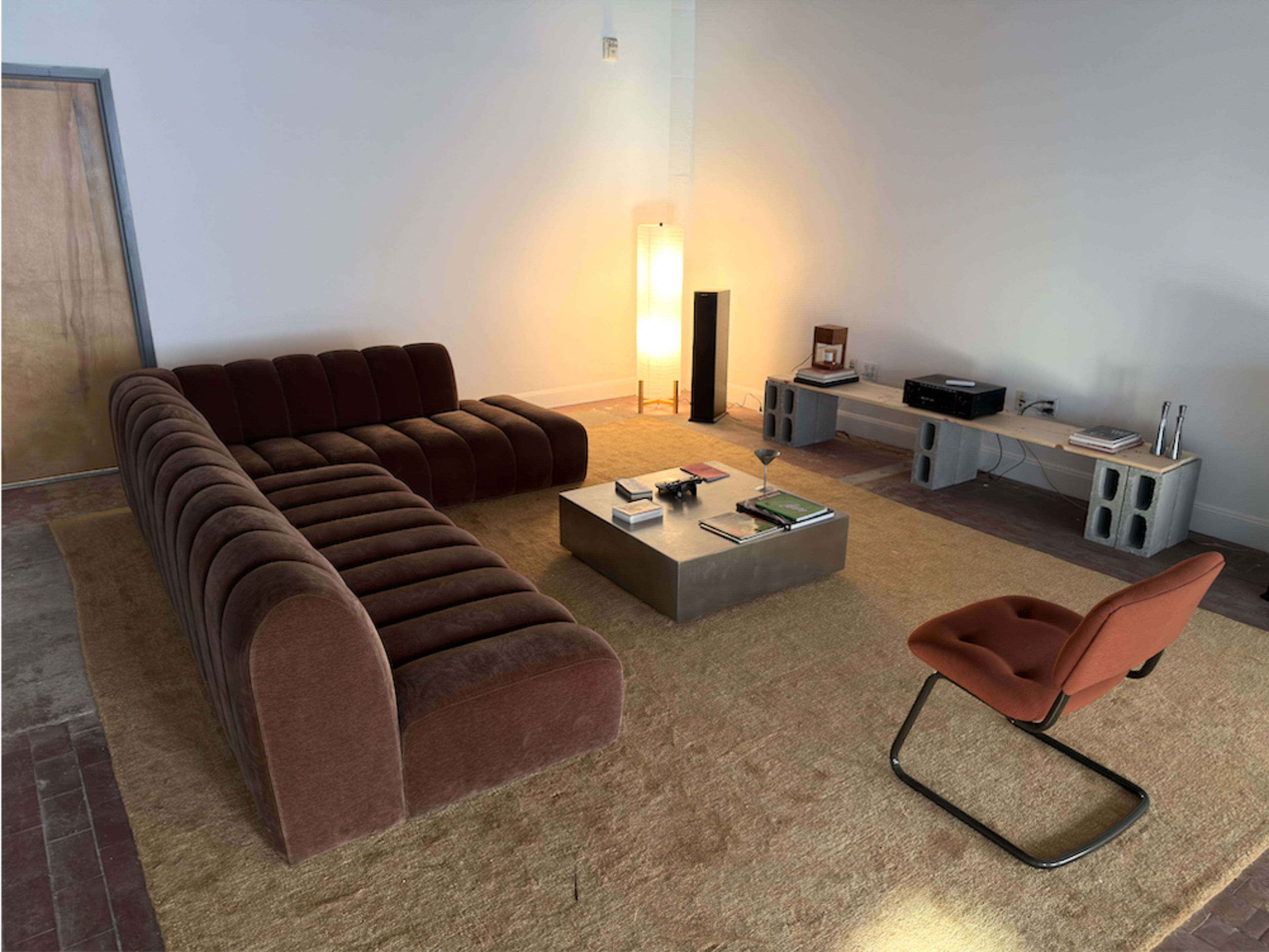 The image shows a modern living room with a brown sectional sofa, an orange accent chair, a low coffee table, and a tall lamp, all placed on a large beige area rug.