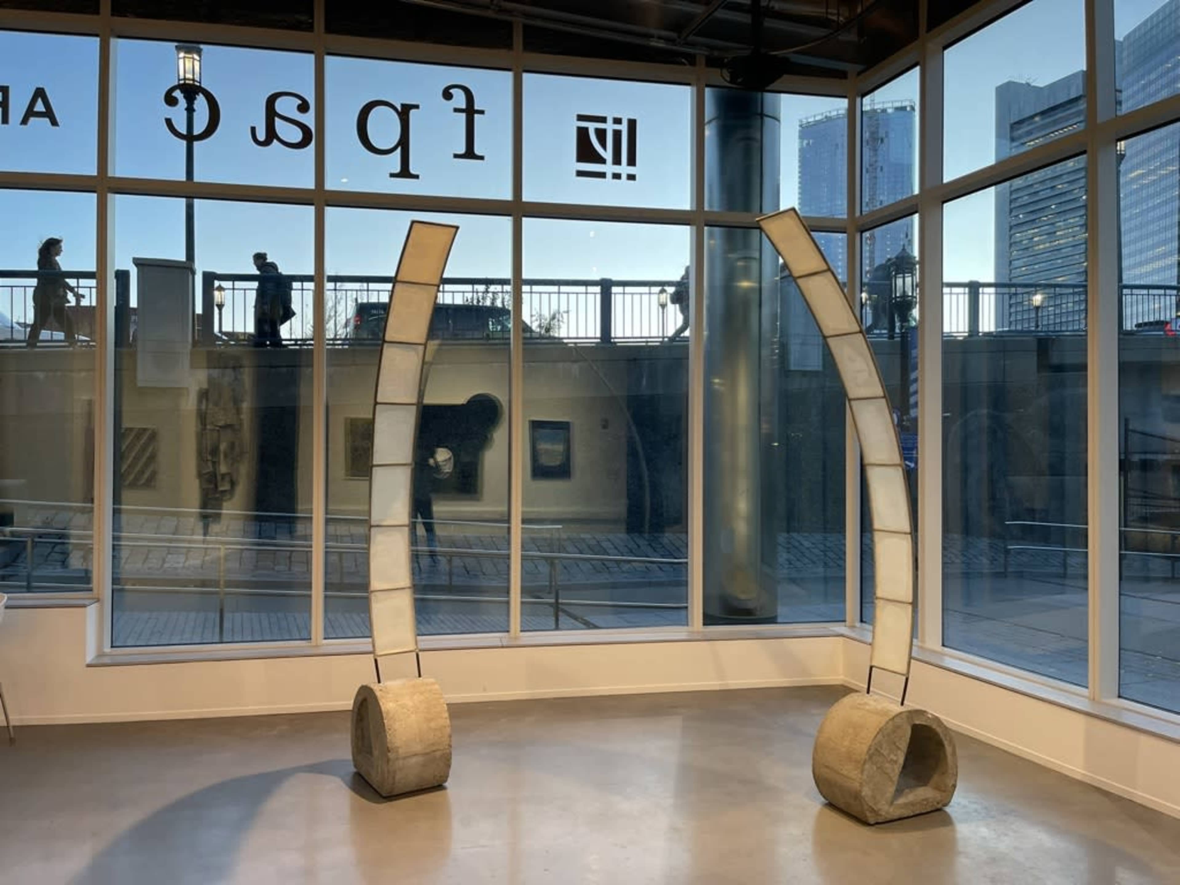 The image shows two curved sculptures made of concrete positioned inside a modern gallery with large windows overlooking a cityscape.